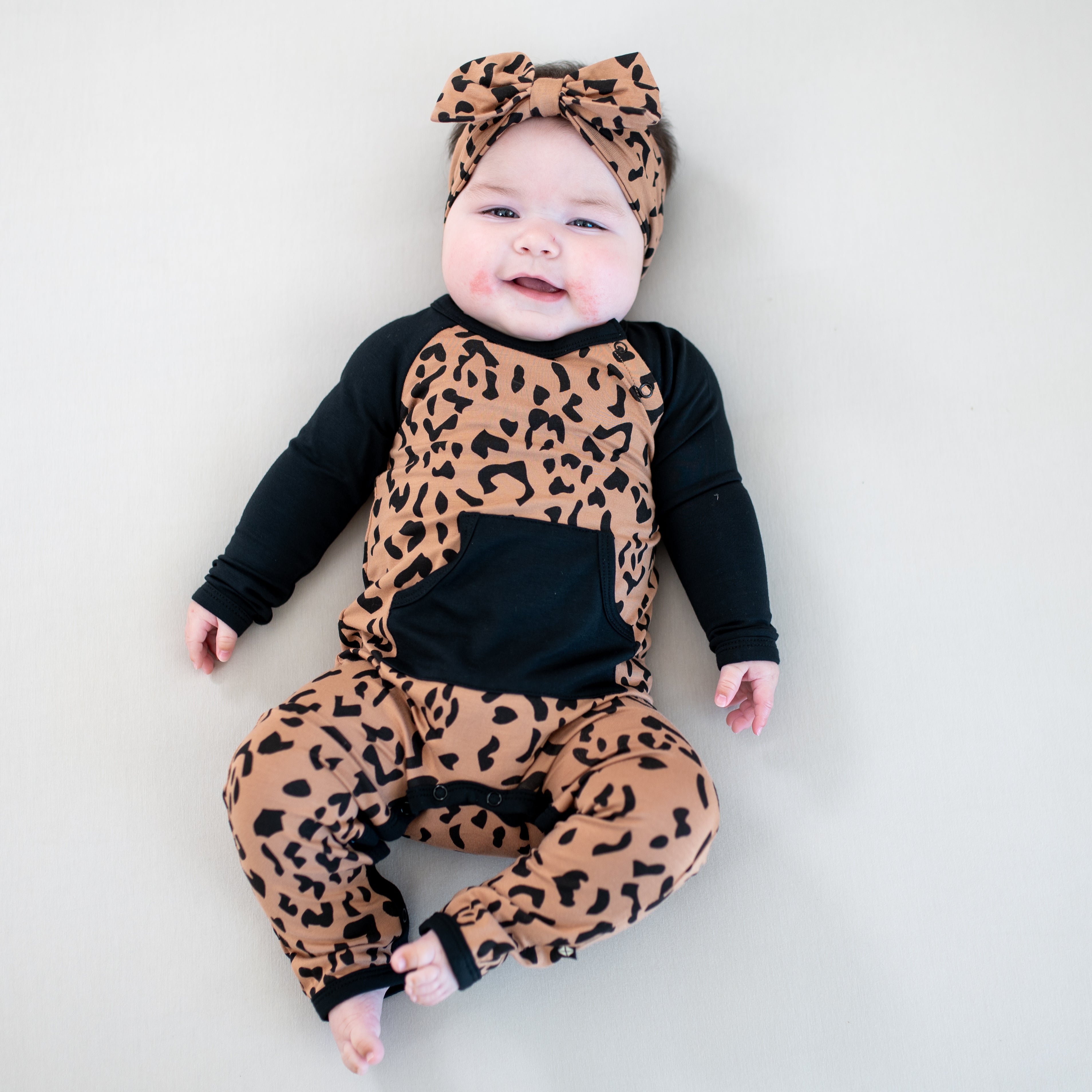 Smiling infant wearing the Longall in Latte Leopard paired with matching bow headband laying on a light colored background