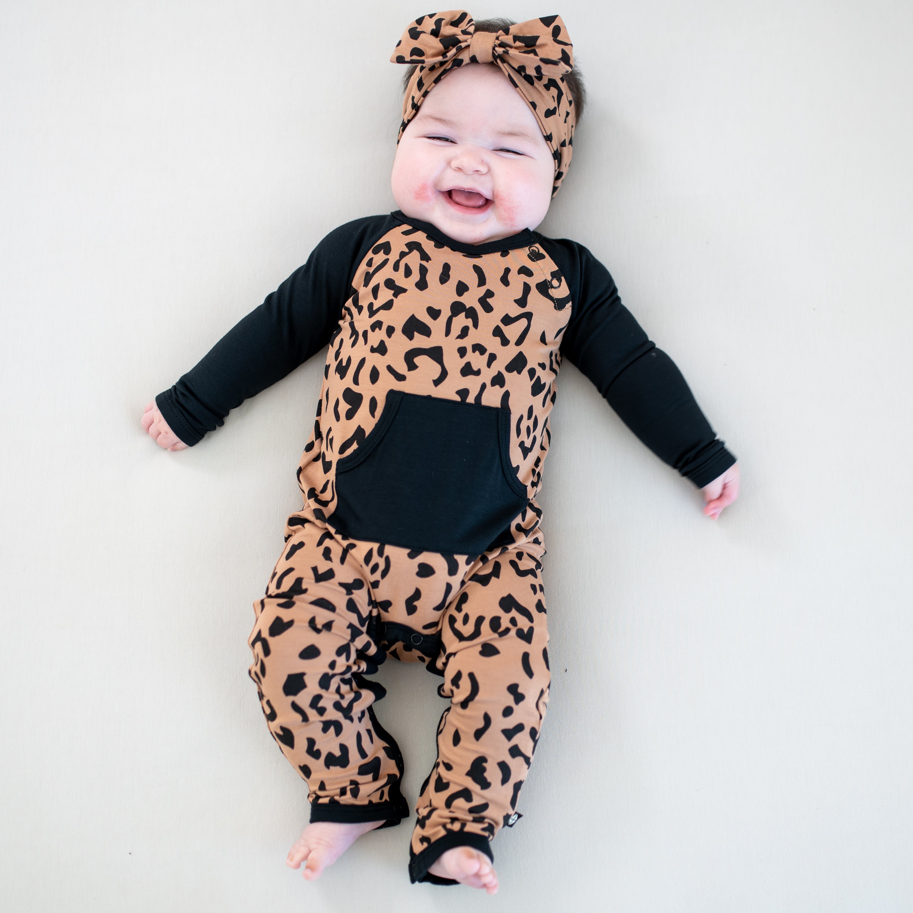 Smiling infant wearing the Longall in Latte Leopard paired with matching bow headband on a light colored background