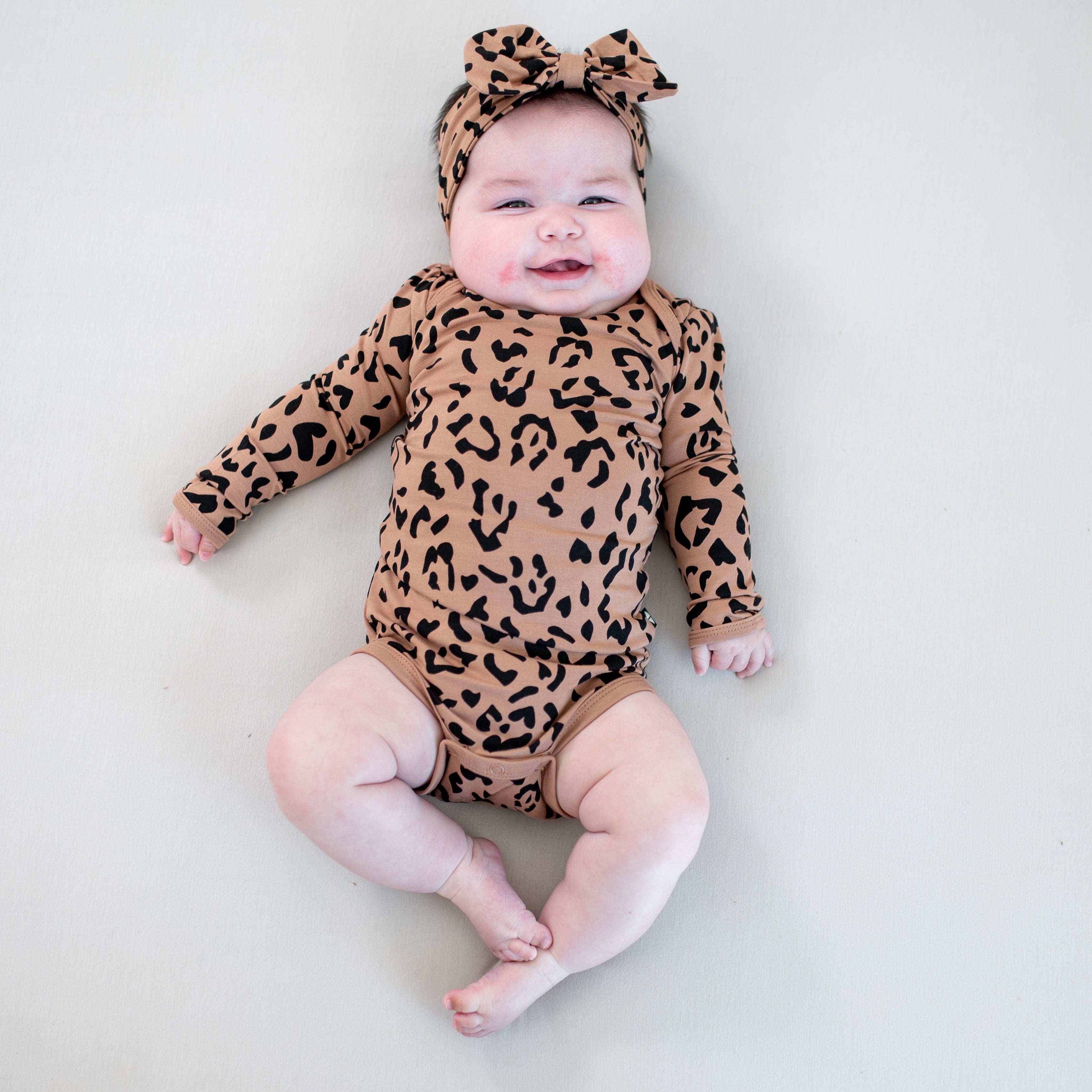 Smiling infant laying on a light colored blanket wearing the Long Sleeve Bodysuit in Latte Leopard with matching bow headband