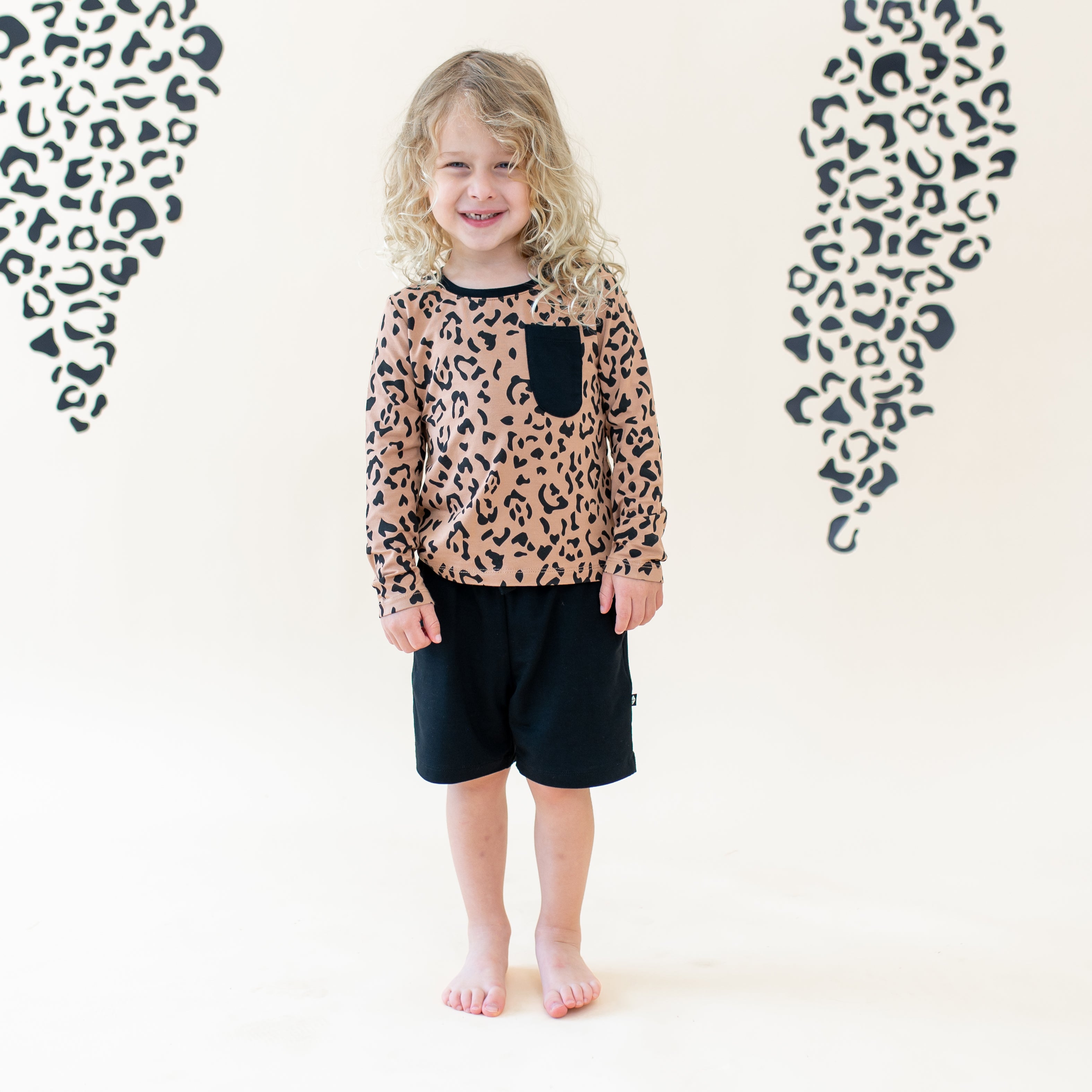 Child model standing wearing the Long Sleeve Toddler Crew Neck Tee in Latte Leopard paired with Midnight Drawstring shorts in front of a leopard print backdrop