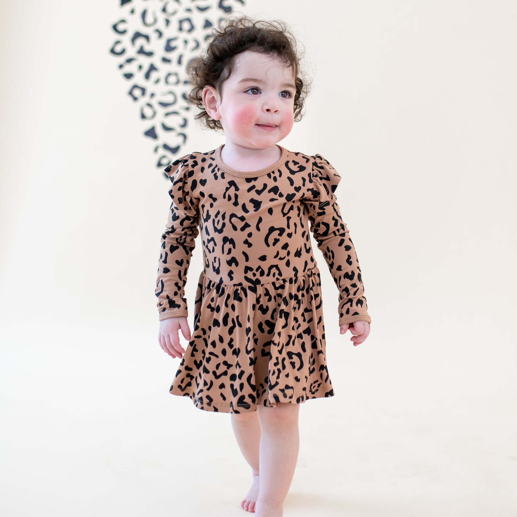 Toddler girl walking wearing the Long Sleeve Twirl Bodysuit Dress in Latte Leopard in front of a light leopard print backdrop