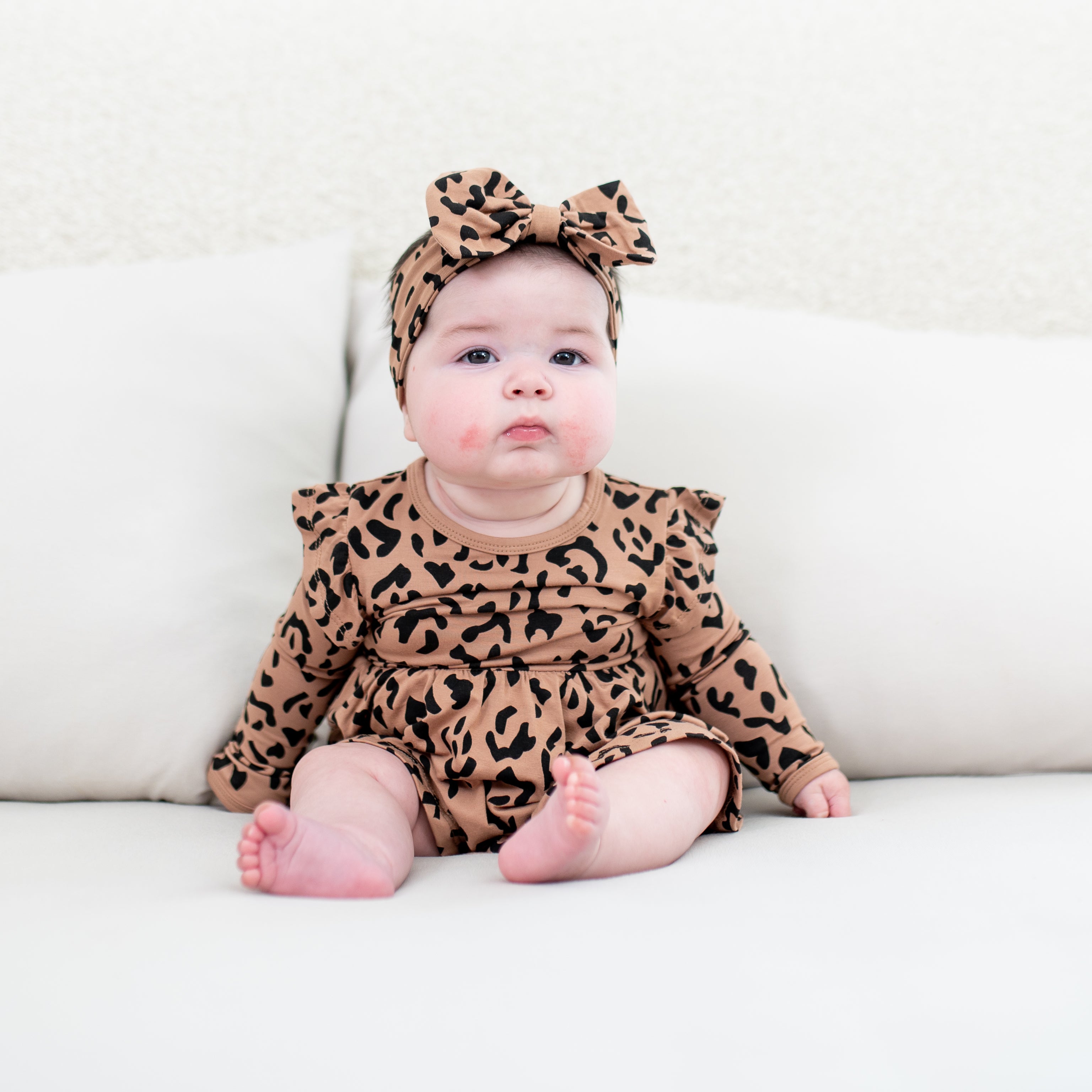 Infant sitting in a bed wearing the Long Sleeve Twirl Bodysuit Dress in Latte Leopard with a matching bow headband