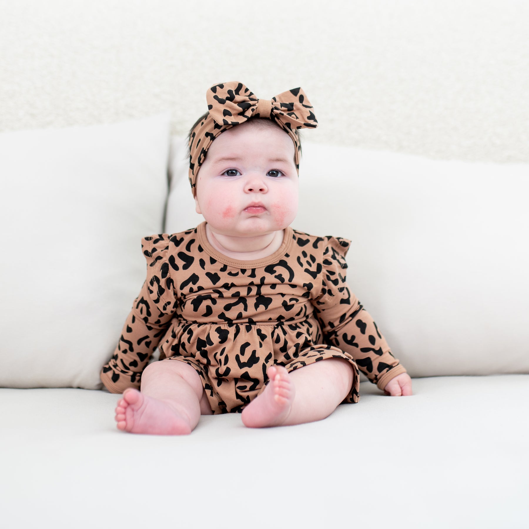 Infant sitting in a bed wearing the Long Sleeve Twirl Bodysuit Dress in Latte Leopard with a matching bow headband