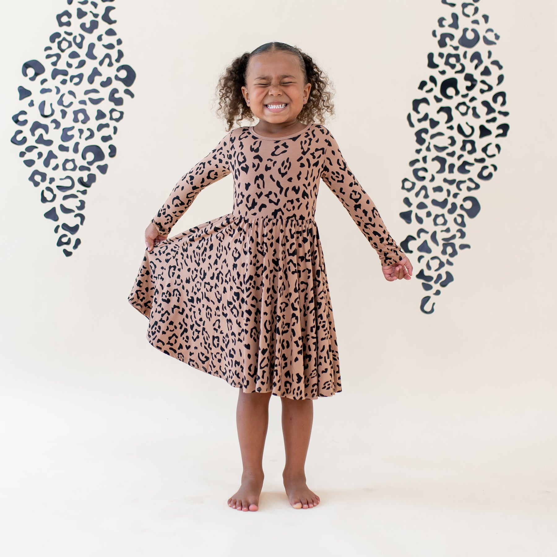 Young girl smiling holding the side of her Long Sleeve Twirl Dress in Latte Leopard out in front of a light colored leopard print backdrop