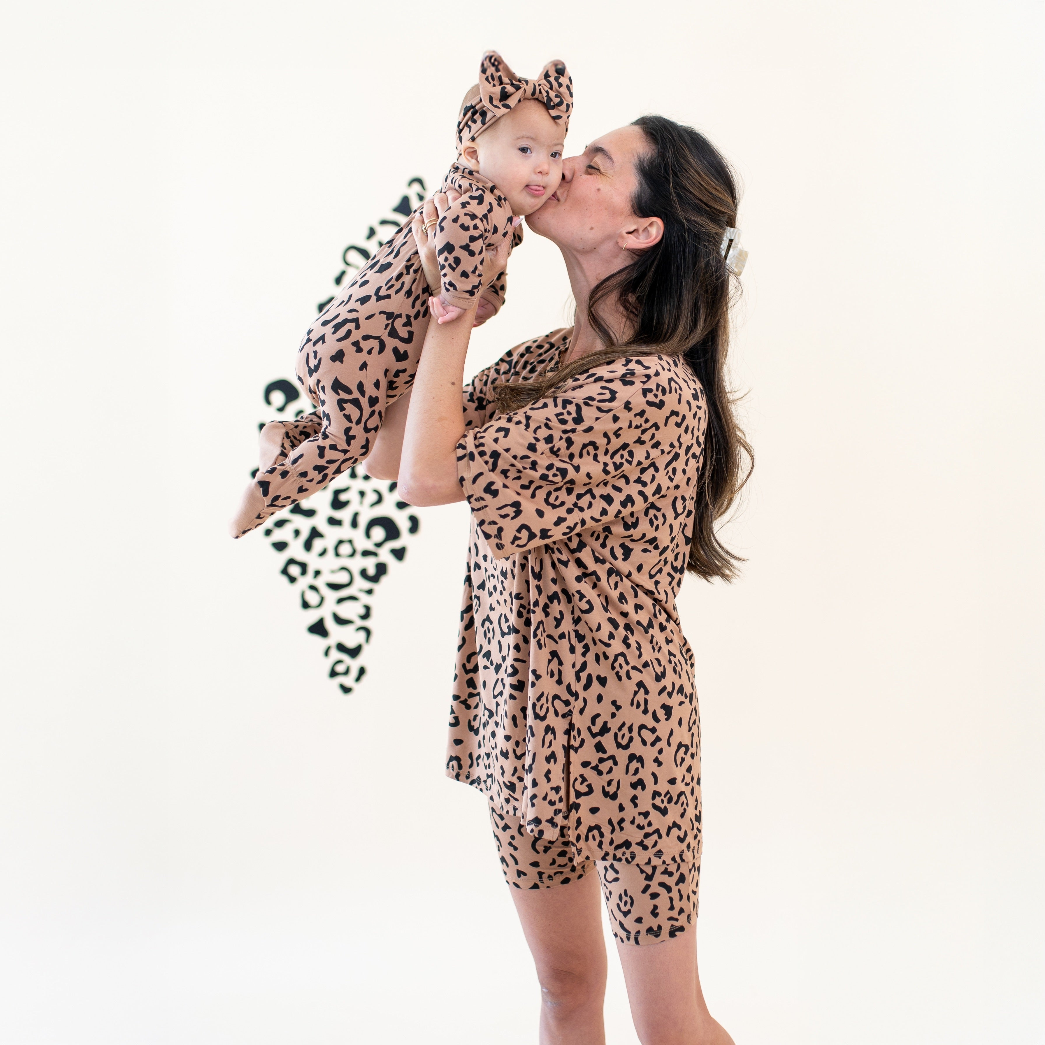 Mother holding infant in the air kissing the baby while baby has her tongue sticking out. Mother wearing the Women’s Biker Short Set in Latte Leopard and infant is matching in the zippered romper and bow headband