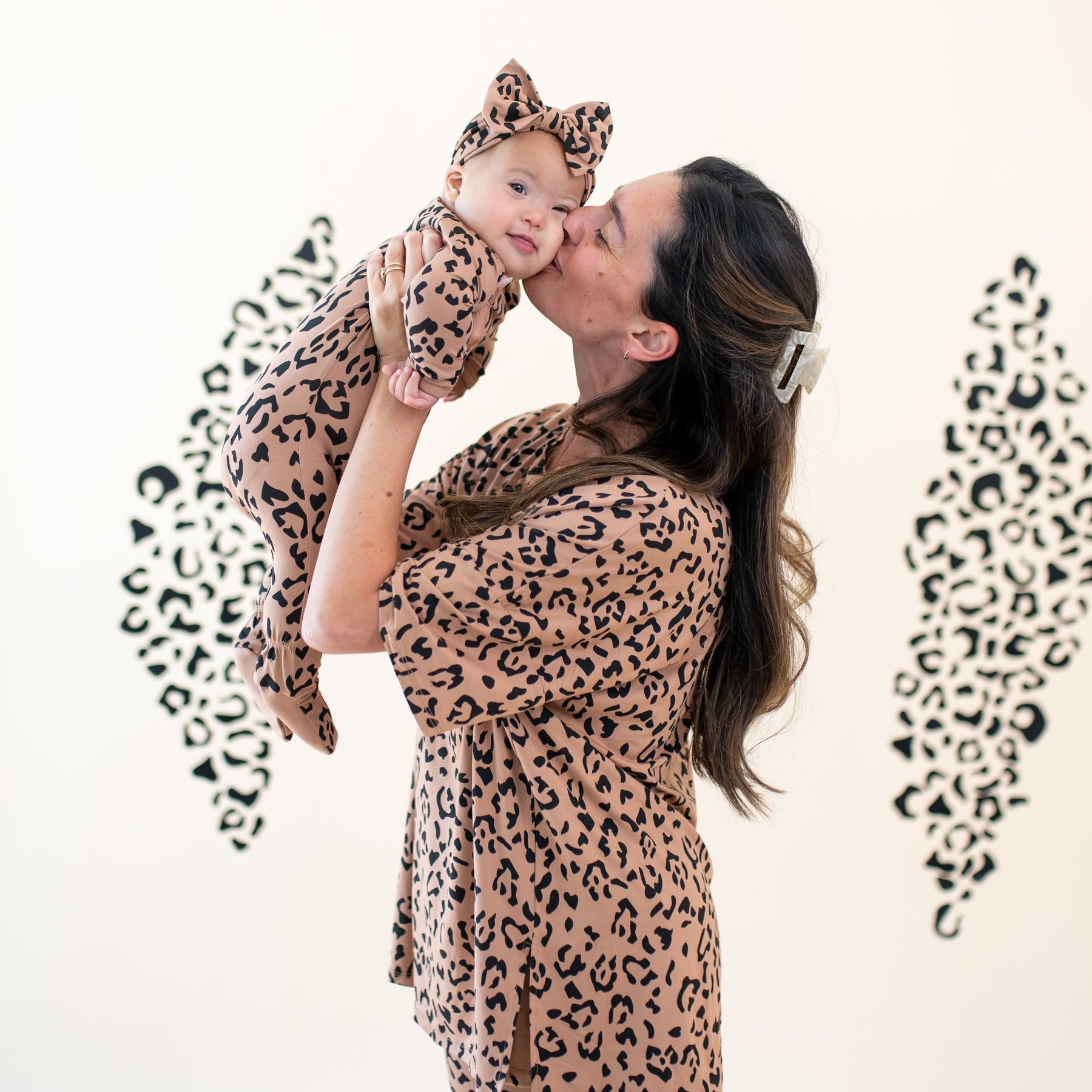 Mother wearing the Women's Biker Short Set in Latte Leopard holding her daughter in the air kissing her daughter on the cheek who is matching in the Bow Headband in Latte Leopard and matching zippered footie