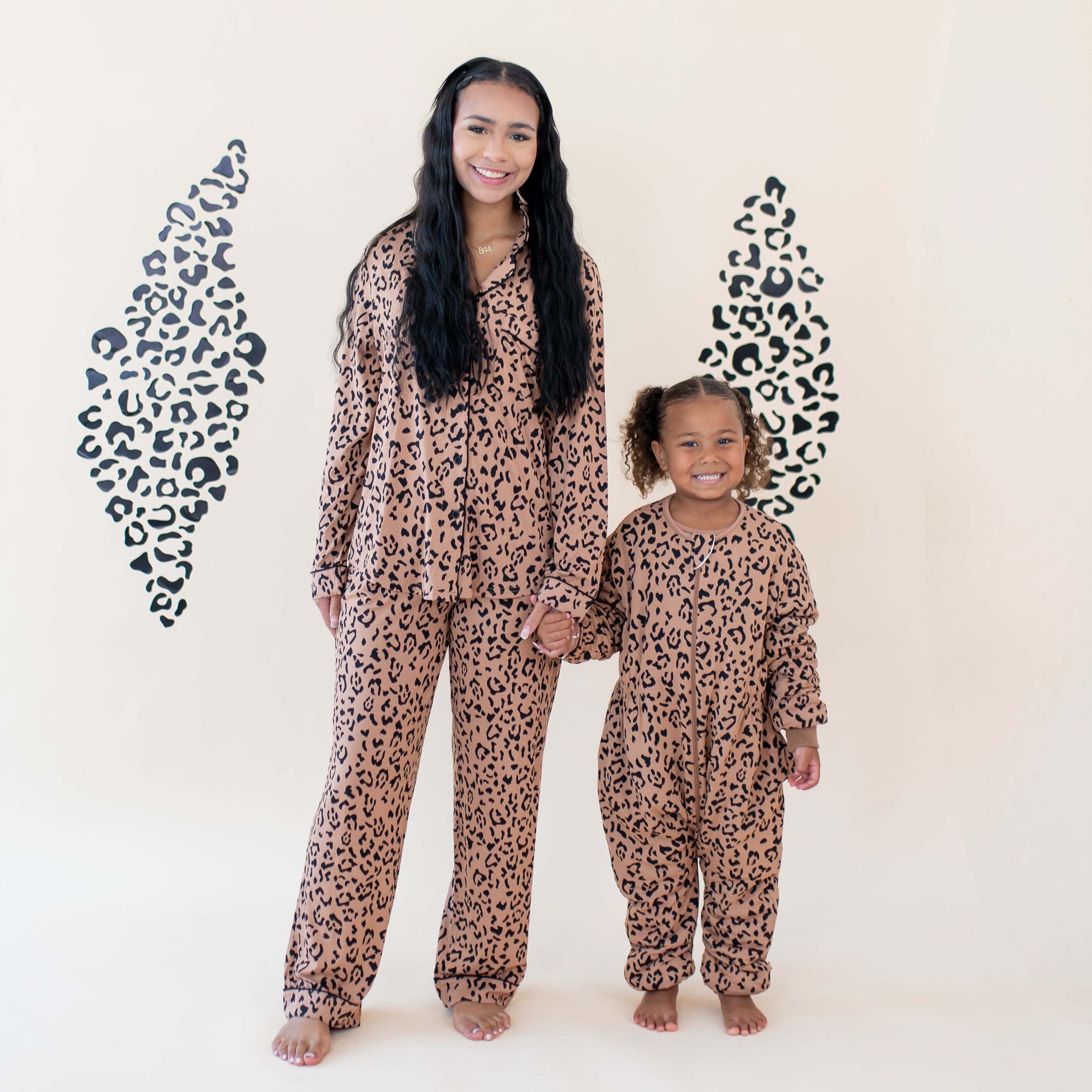 Mother and daughter standing in front of a neutral leopard backdrop holding hands wearing the Cozy Playsuit in Latte Leopard and matching Women's Long Sleeve Pajama set