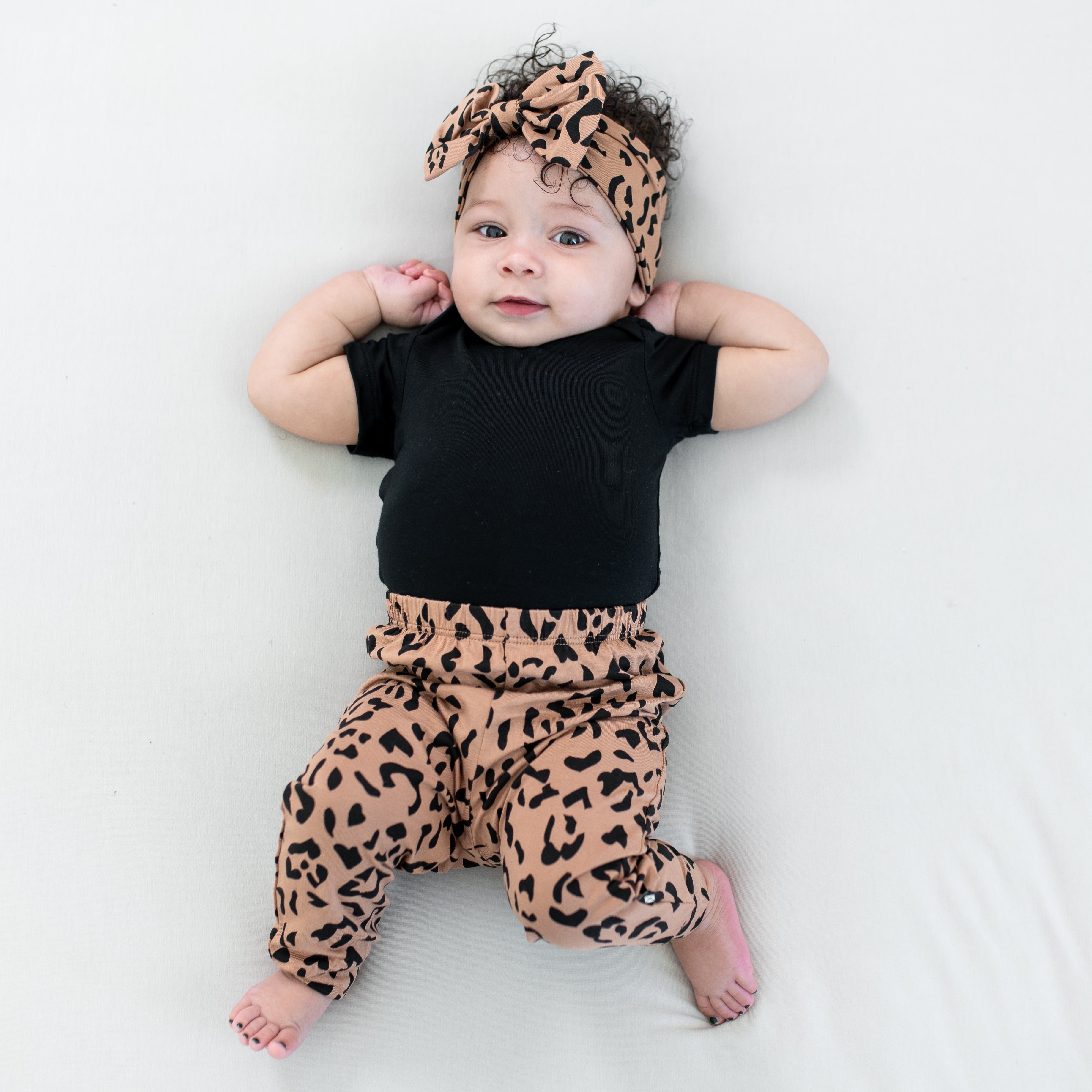 Infant wearing the Pant in Latte Leopard paired with a matching bow headband and midnight short sleeve bodysuit laying on a light neutral blanket