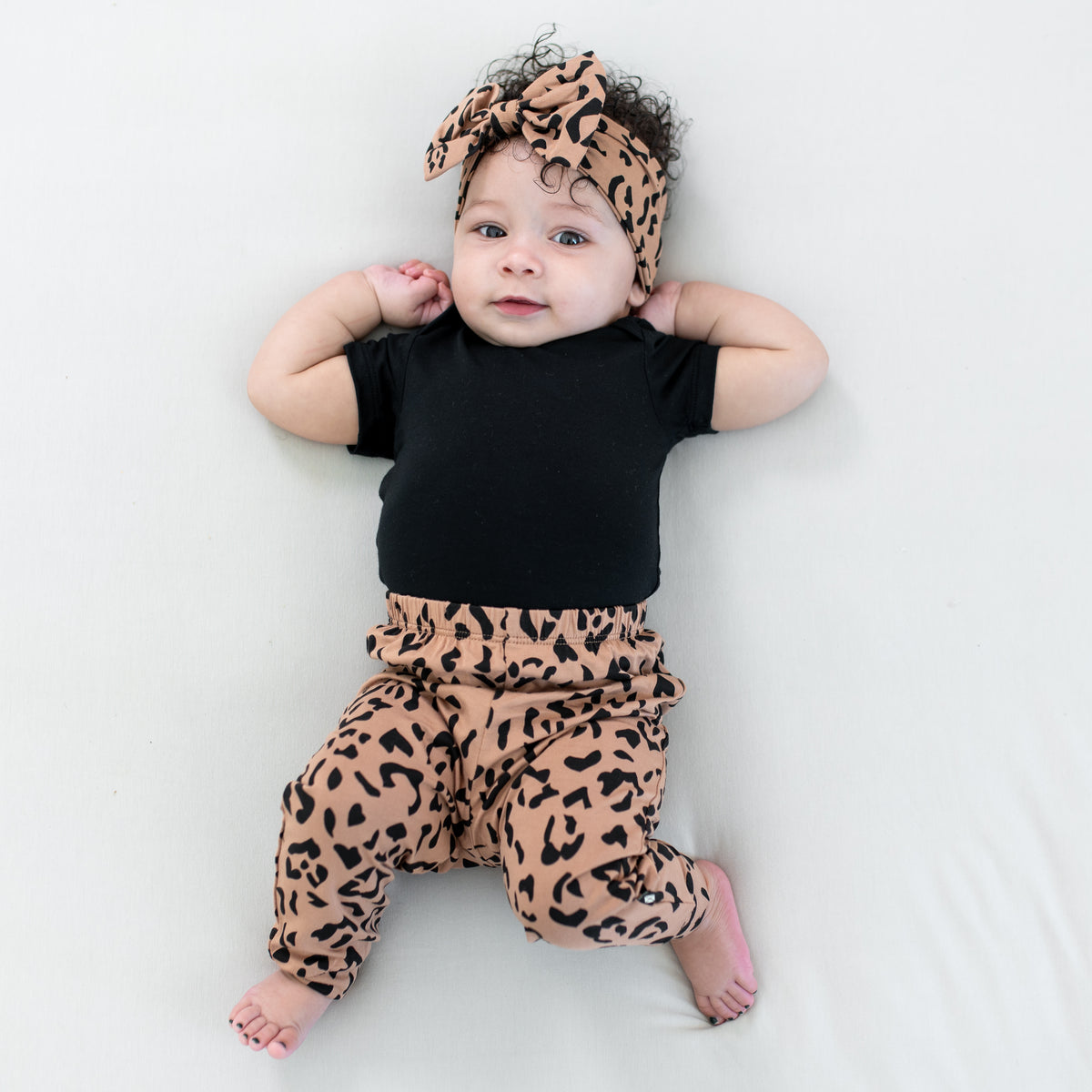 Infant wearing the Pant in Latte Leopard paired with a matching bow headband and midnight short sleeve bodysuit laying on a light neutral blanket