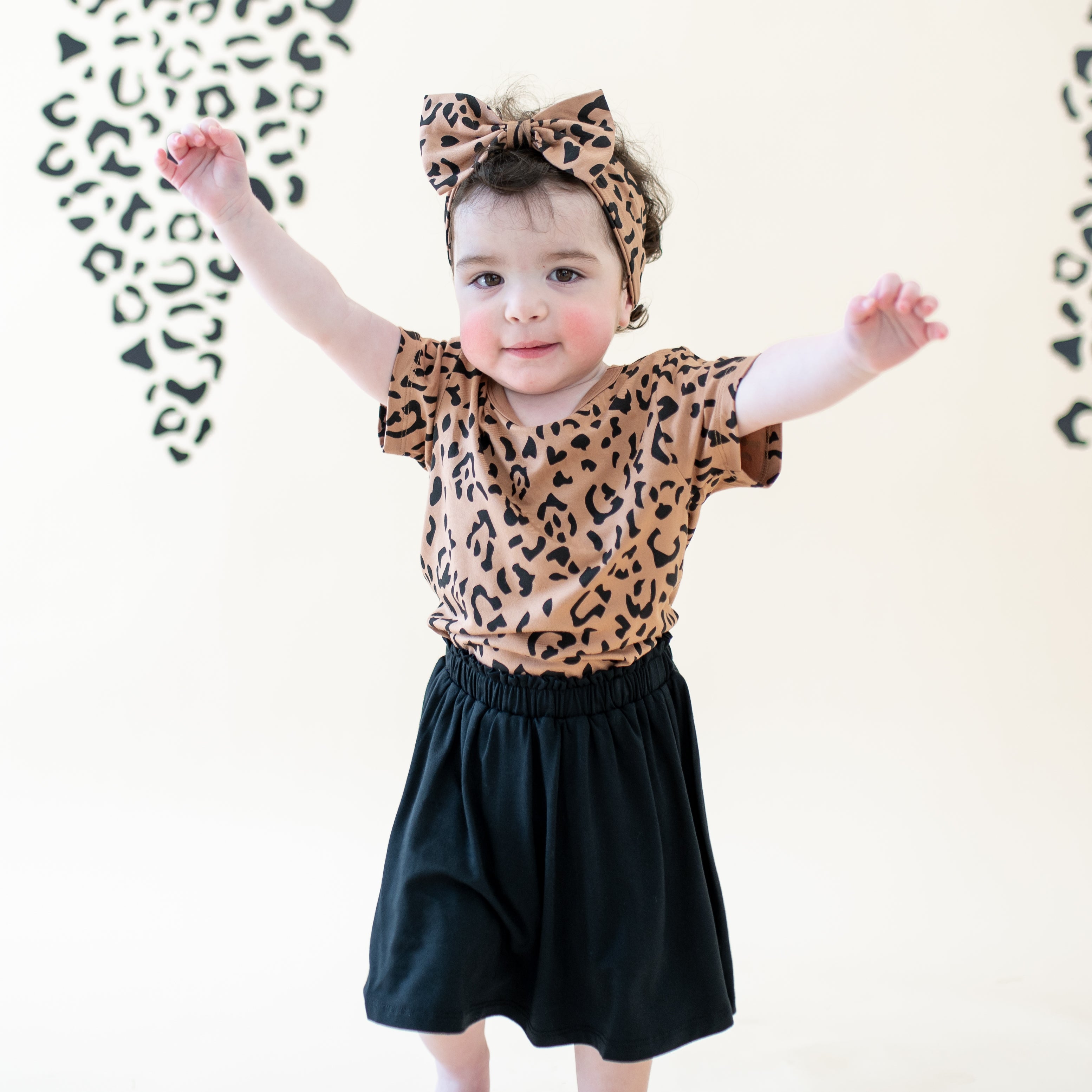 Toddler with her hands in the air wearing the Toddler Basic Tee in Latte Leopard paired with matching bow headband and Midnight skort