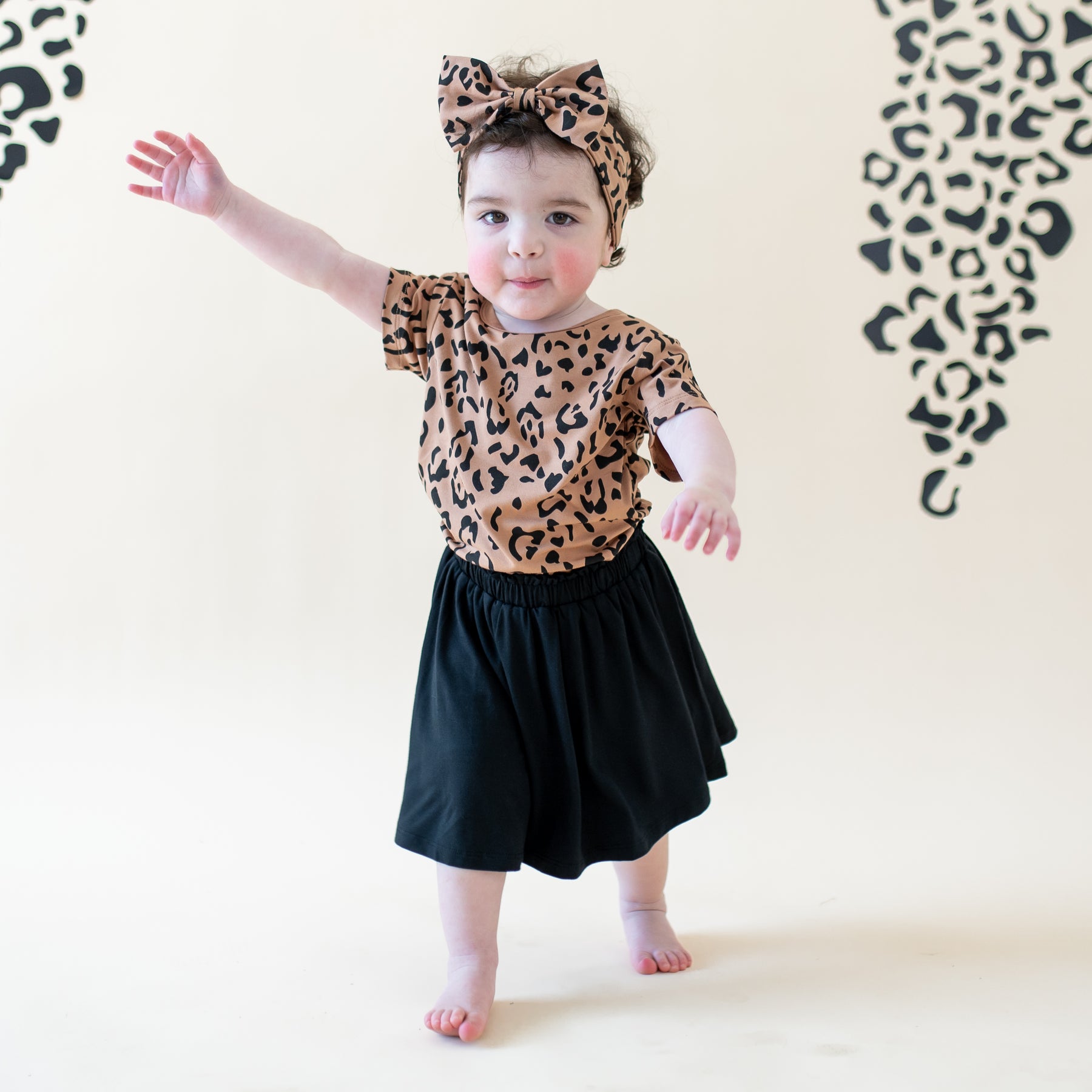 Toddler walking wearing the Toddler Basic Tee in Latte Leopard with matching bow headband and midnight skort