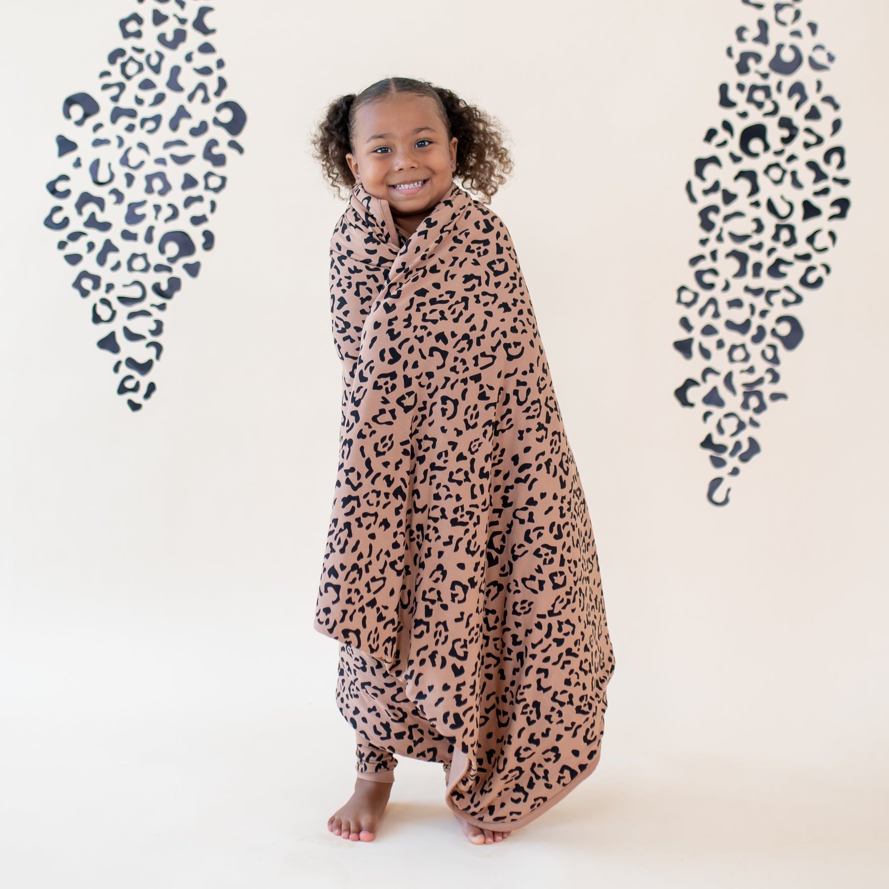 Smiling girl with the Toddler Blanket in Latte Leopard 1.0 wrapped around her in front of a light neutral leopard print back drop