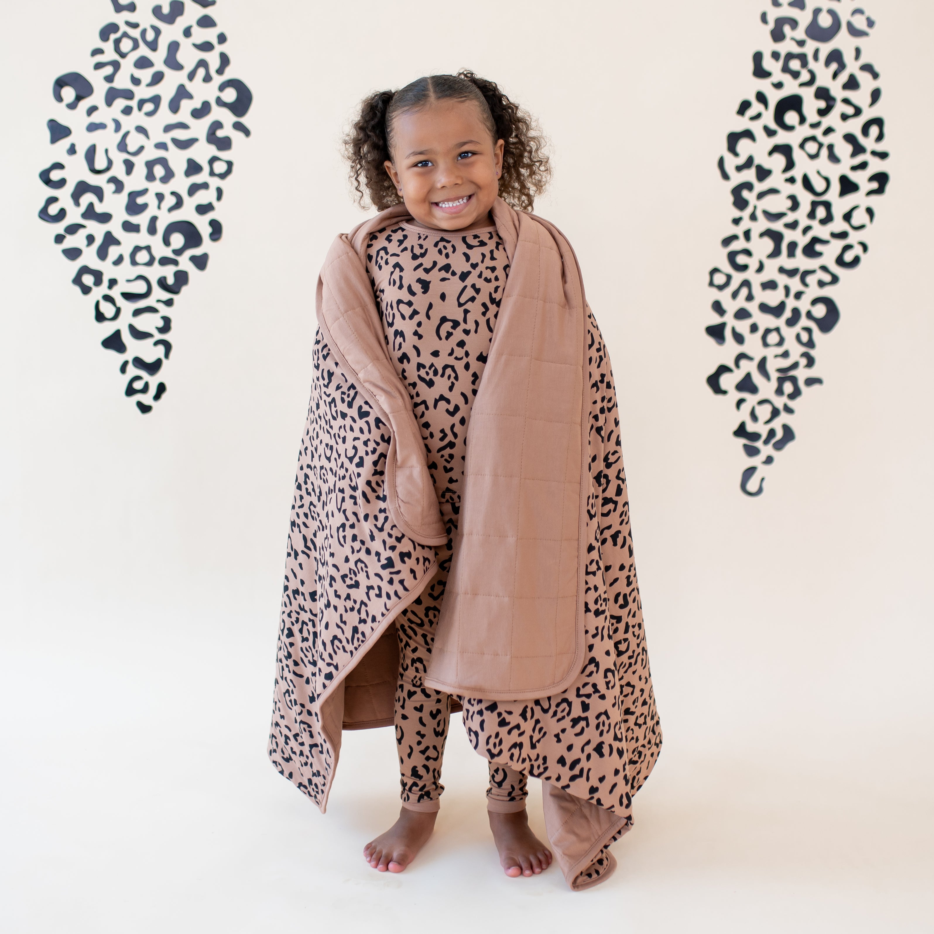 Smiling kid standing with the Toddler Blanket in Latte Leopard 1.0 wrapped around her wearing matching pajamas