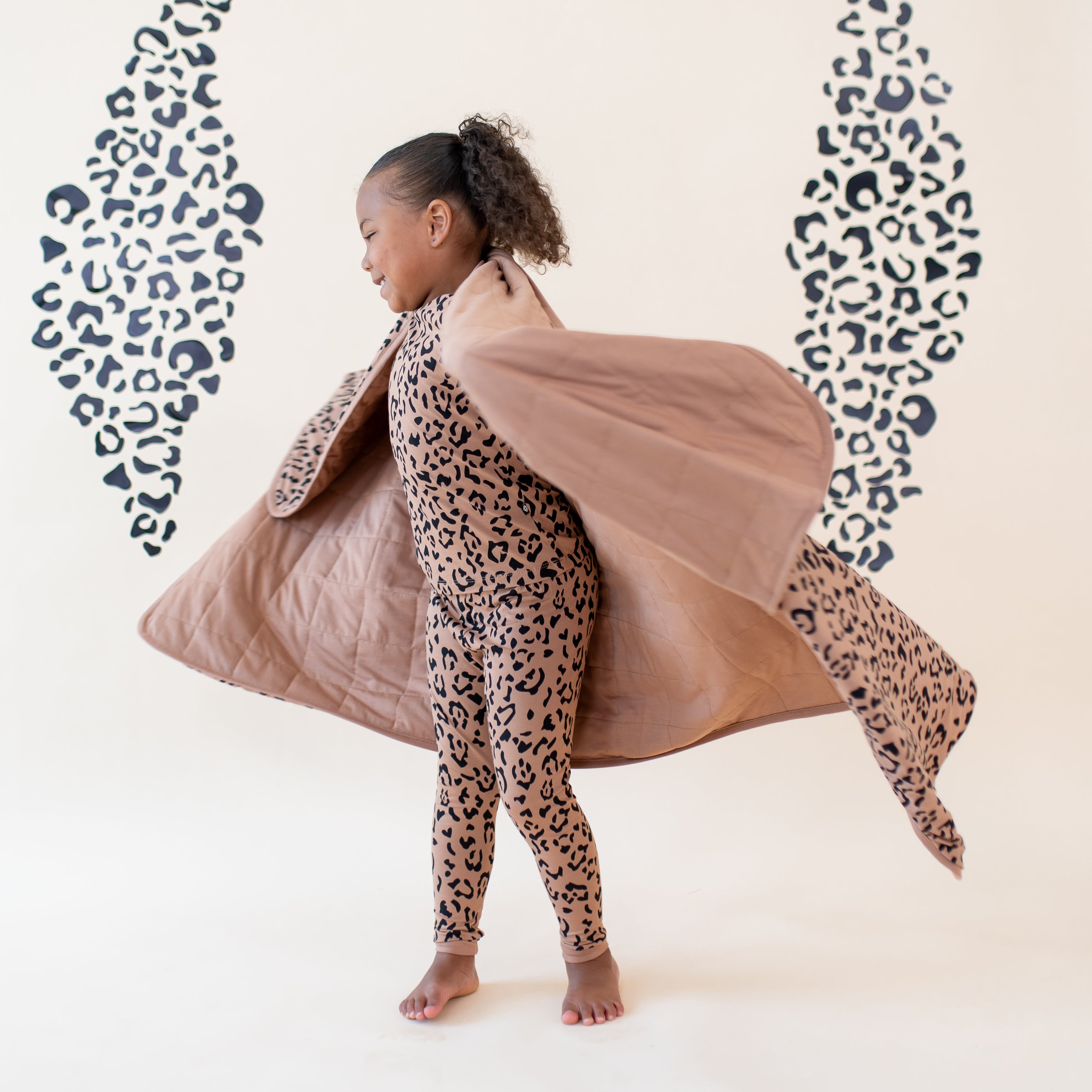 Young girl twirling with the Toddler Blanket in Latte Leopard 1.0 around her shoulders wearing the matching long sleeve toddler pajamas