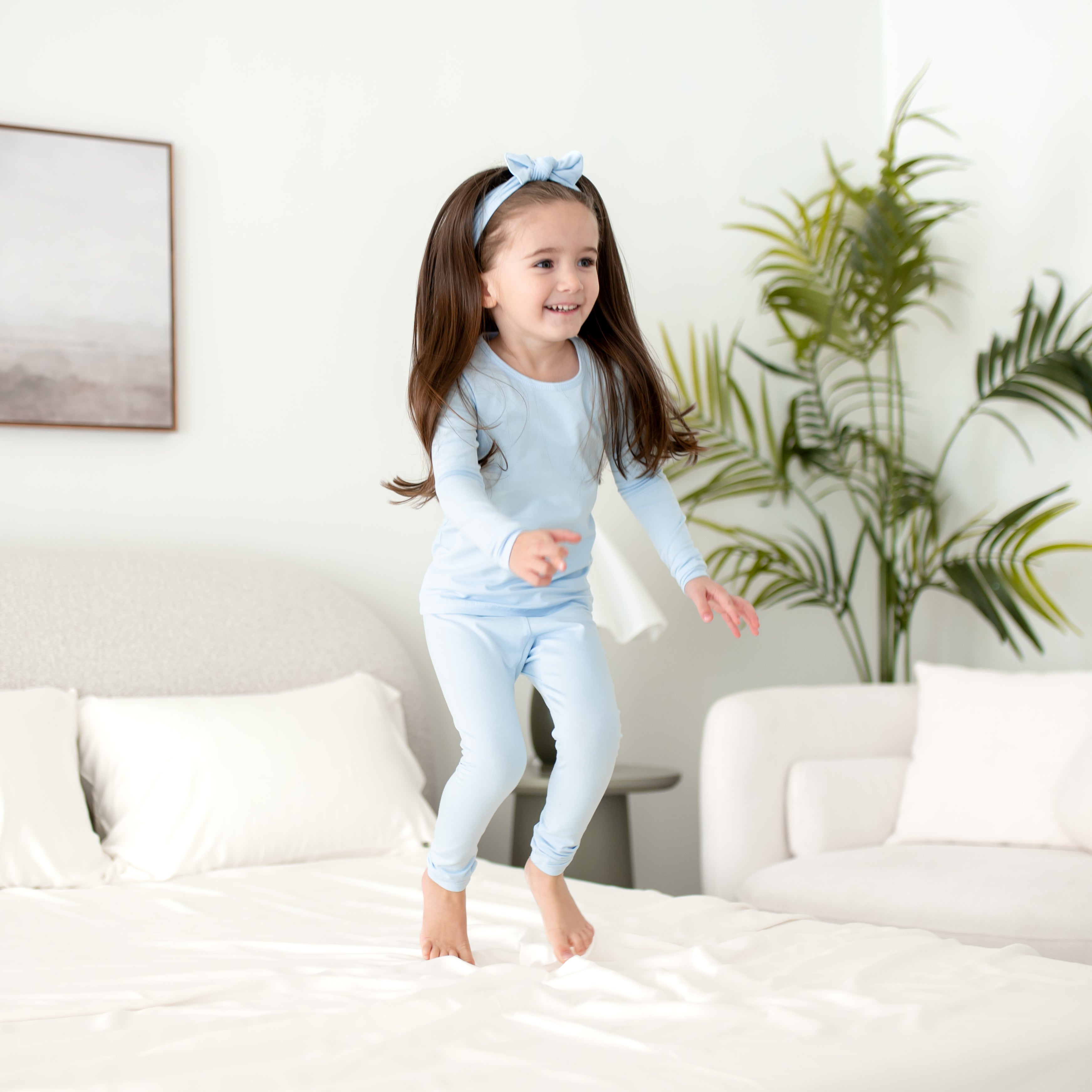 Child in light blue pajamas jumping on a bed in a bright room with a plant in the background