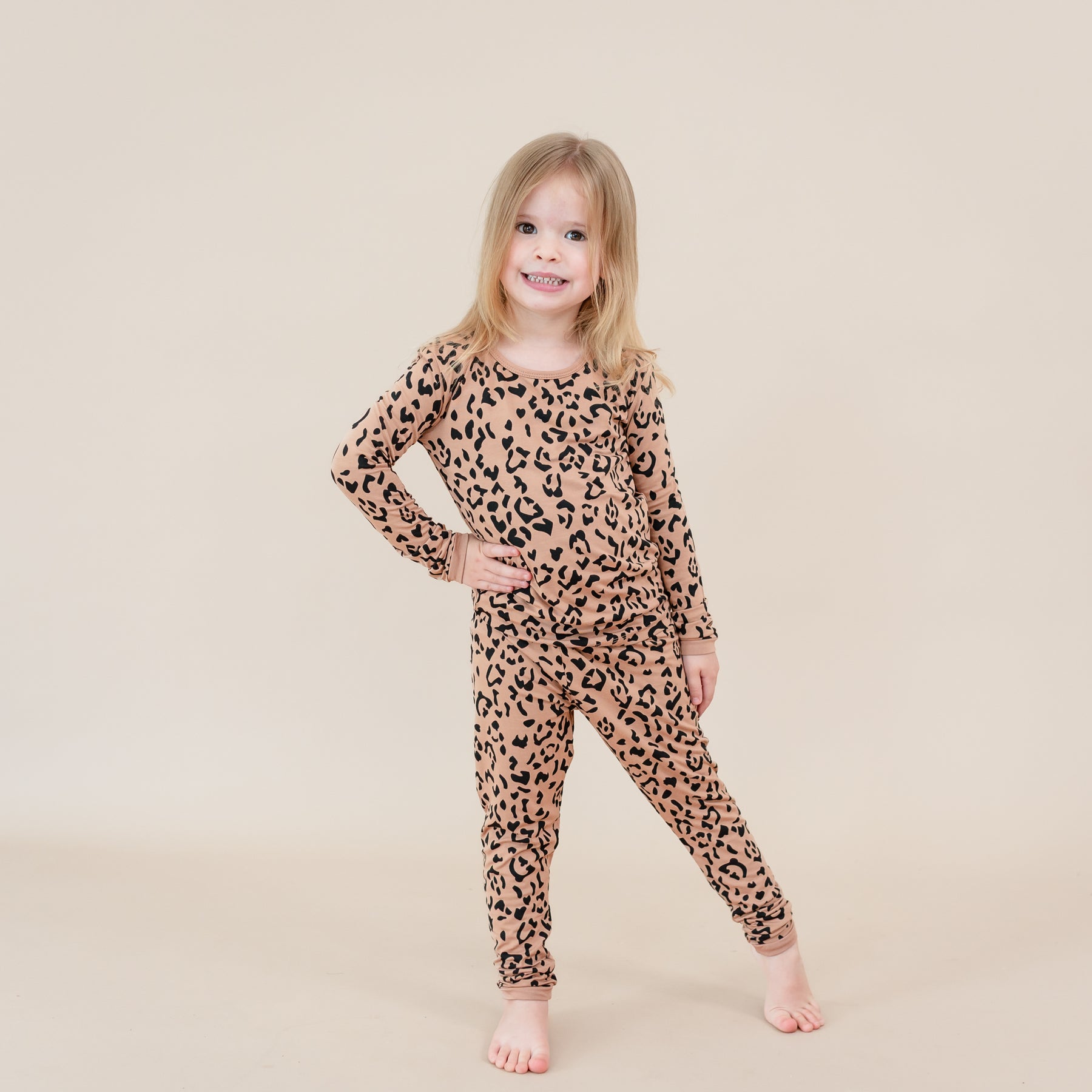 Toddler girl standing with one hand on her hip and one leg off to the side wearing the Long Sleeve Pajamas in Latte Leopard in front of a light beige backdrop