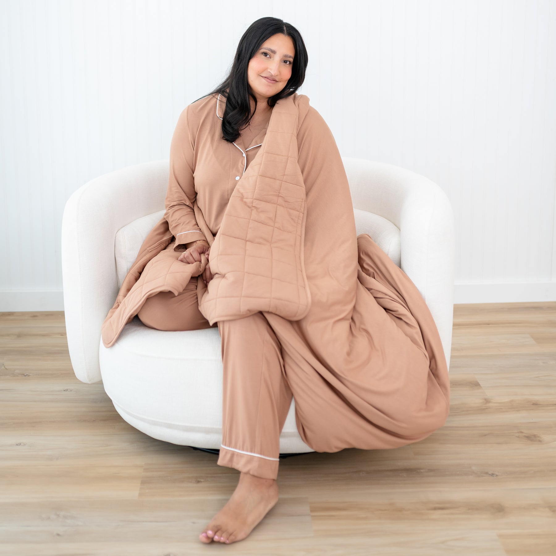 Female model sitting on a couch with the Adult Quilted Blanket in Latte 2.5 wrapped around her