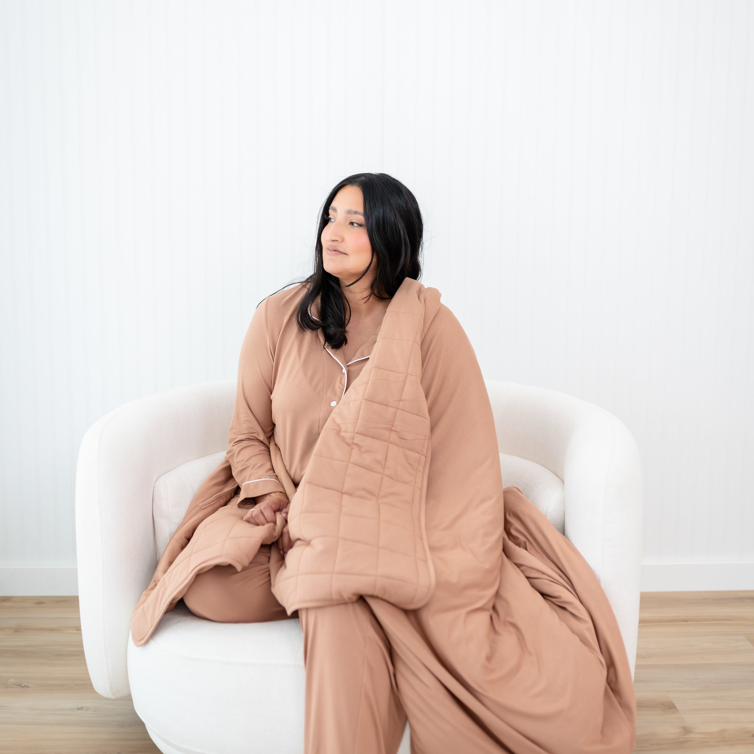 Female sitting on a couch looking off to the side with the Adult Quilted Blanket in Latte 2.5 wrapped around her