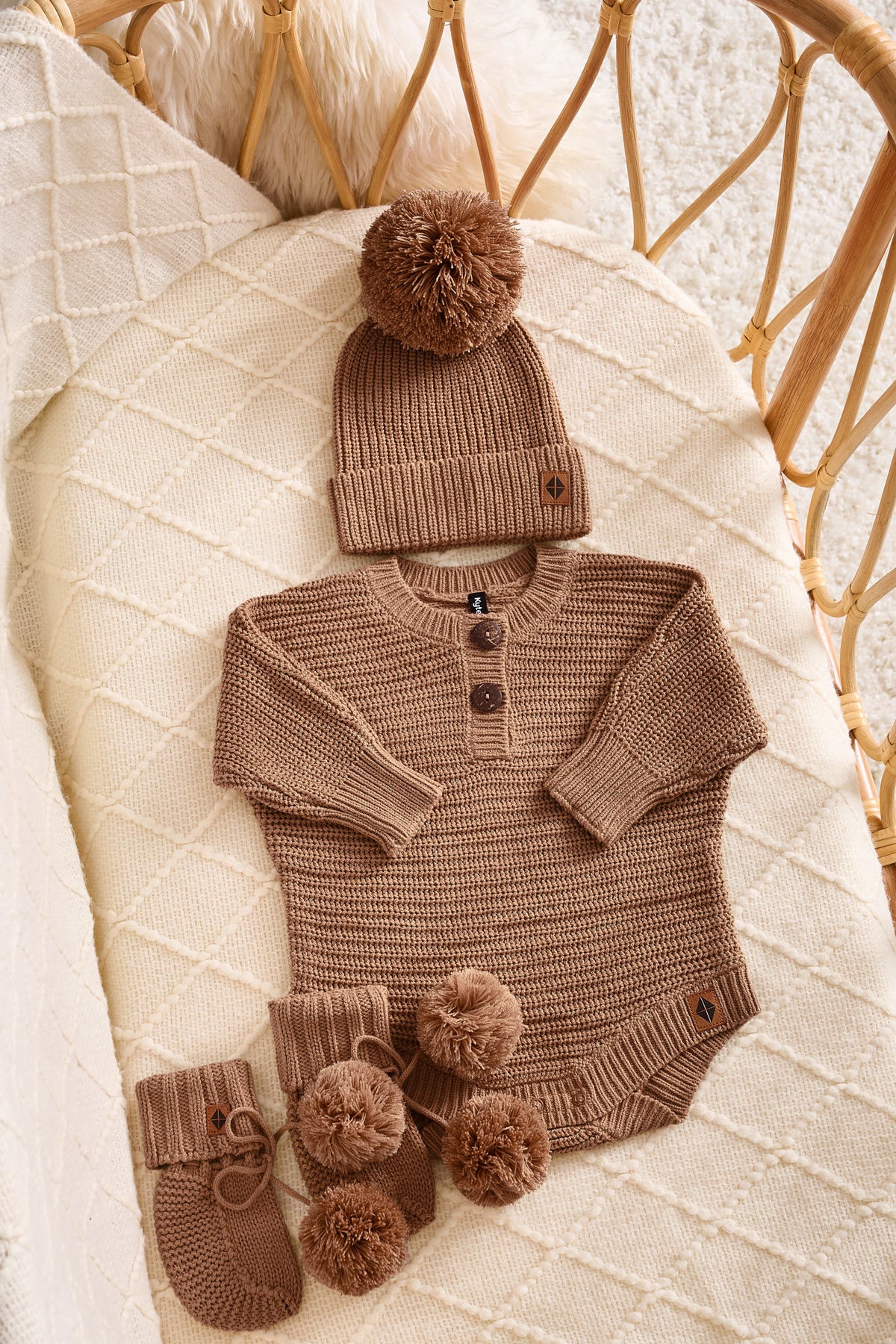 A baby knit outfit set in warm taupe-brown, including a long-sleeve henley bodysuit with three buttons, a matching beanie with a pom-pom, and a pair of booties with pom-poms, arranged on a cream quilted blanket in a rattan bassinet.