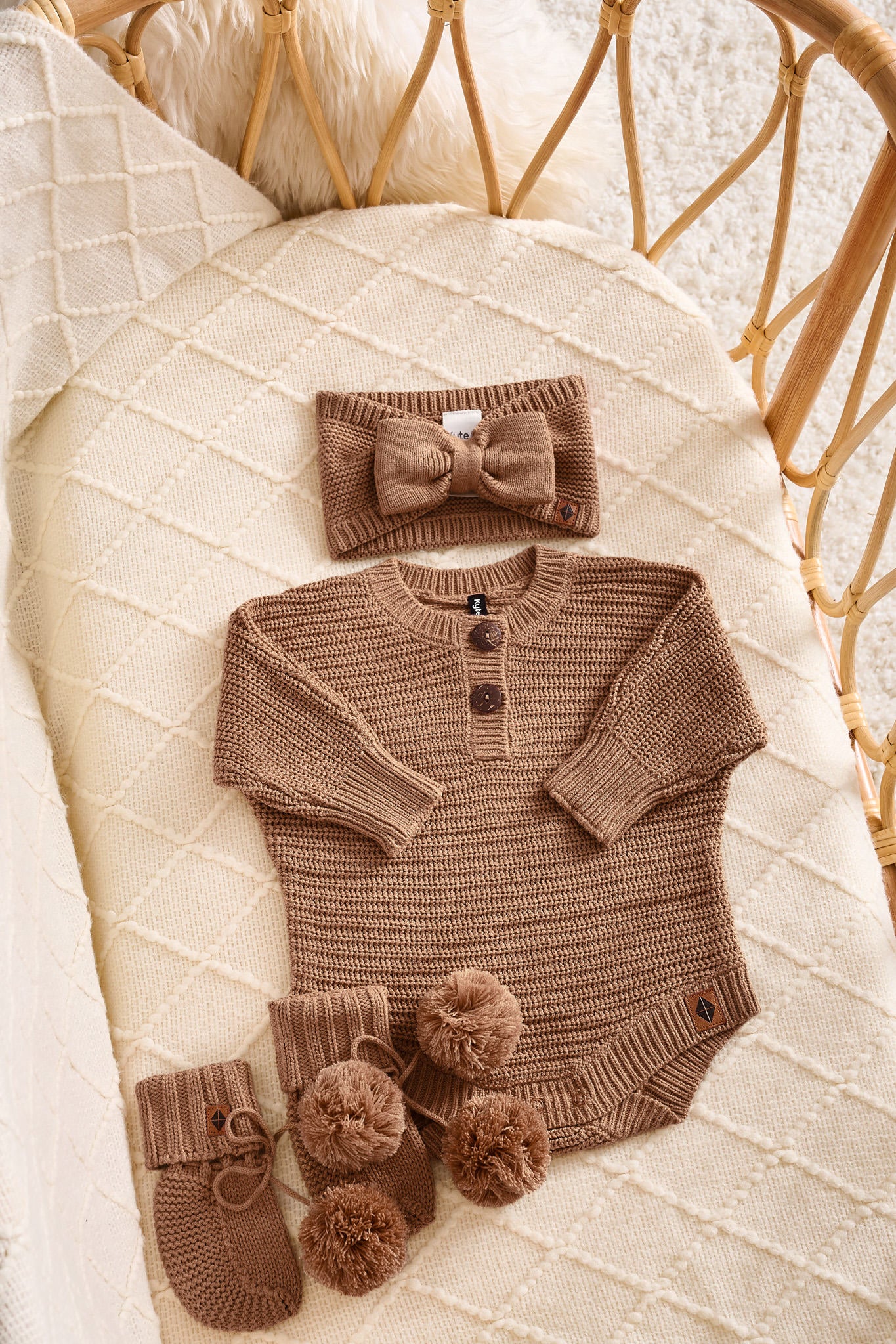 A baby knit outfit set in a warm taupe-brown color, including a long-sleeve henley bodysuit with three buttons, a matching bow headband, and two booties with pom-poms, arranged on a cream quilted blanket inside a rattan bassinet.