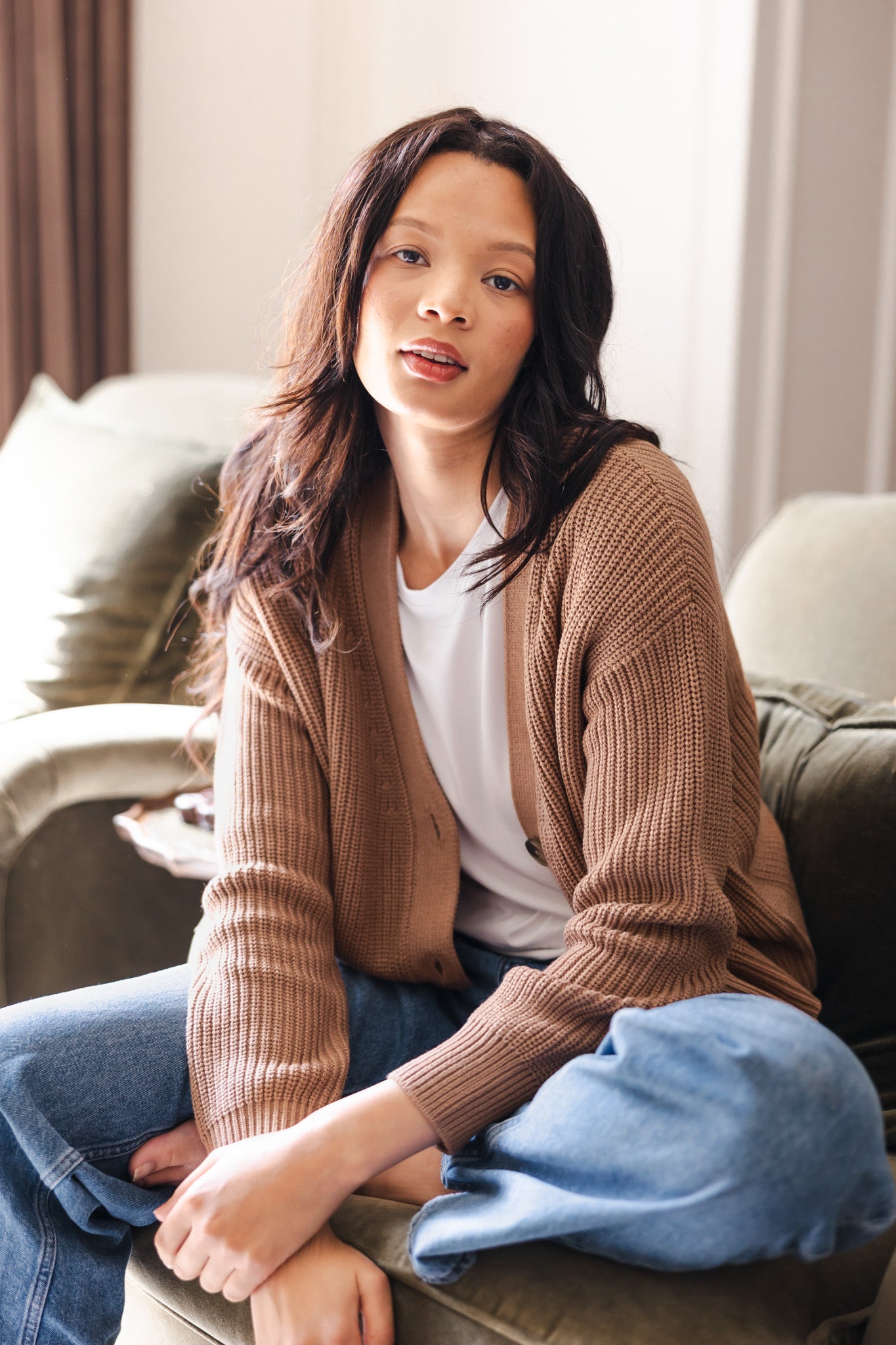 Modal sitting on sofa in Chunky Knit Women's Cropped Cardigan in Latte over white T-shirt and blue jeans