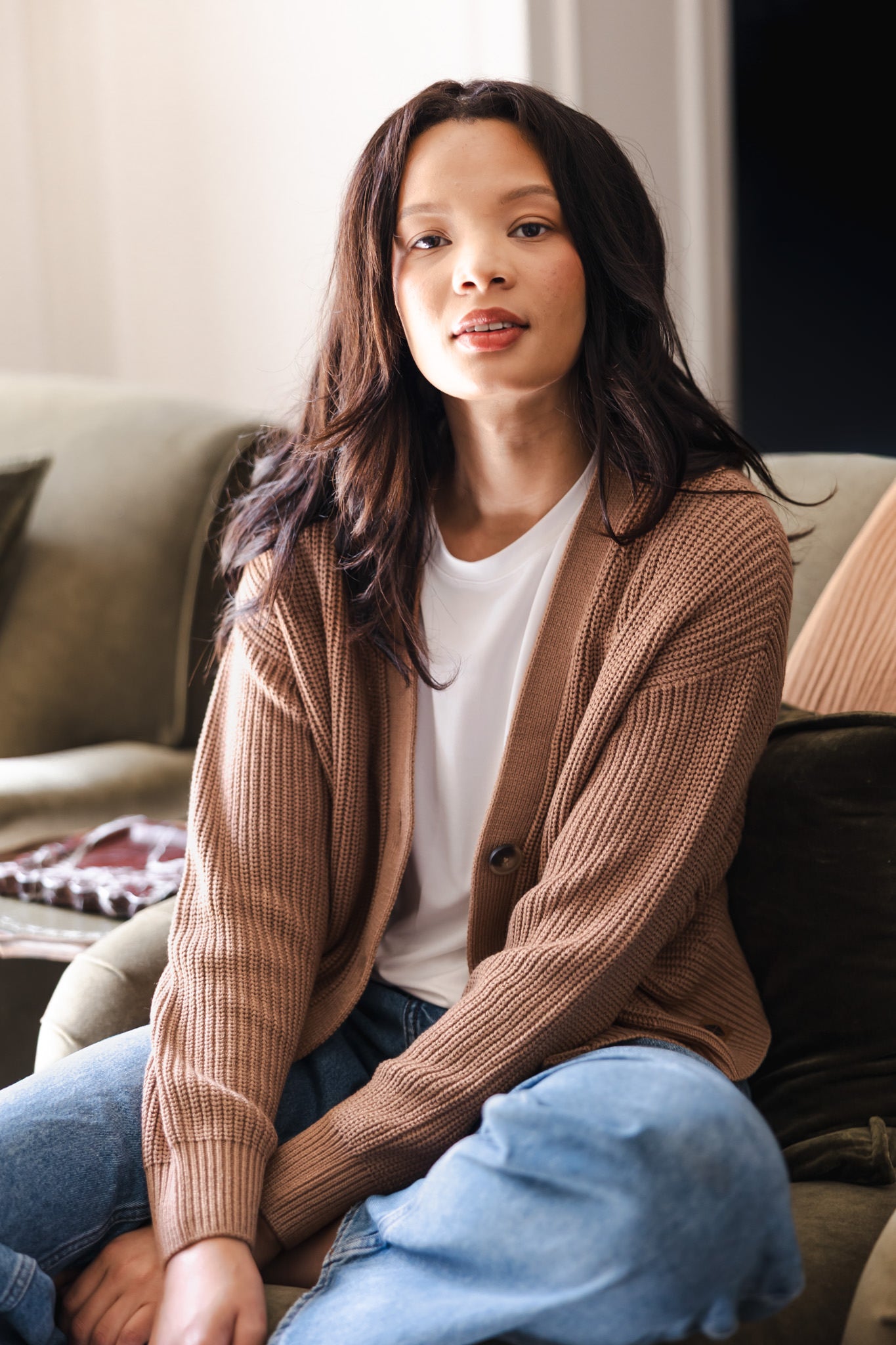 Modal posing on sofa wearing Chunky Knit Women's Cropped Cardigan in Latte over white T-shirt and blue jeans