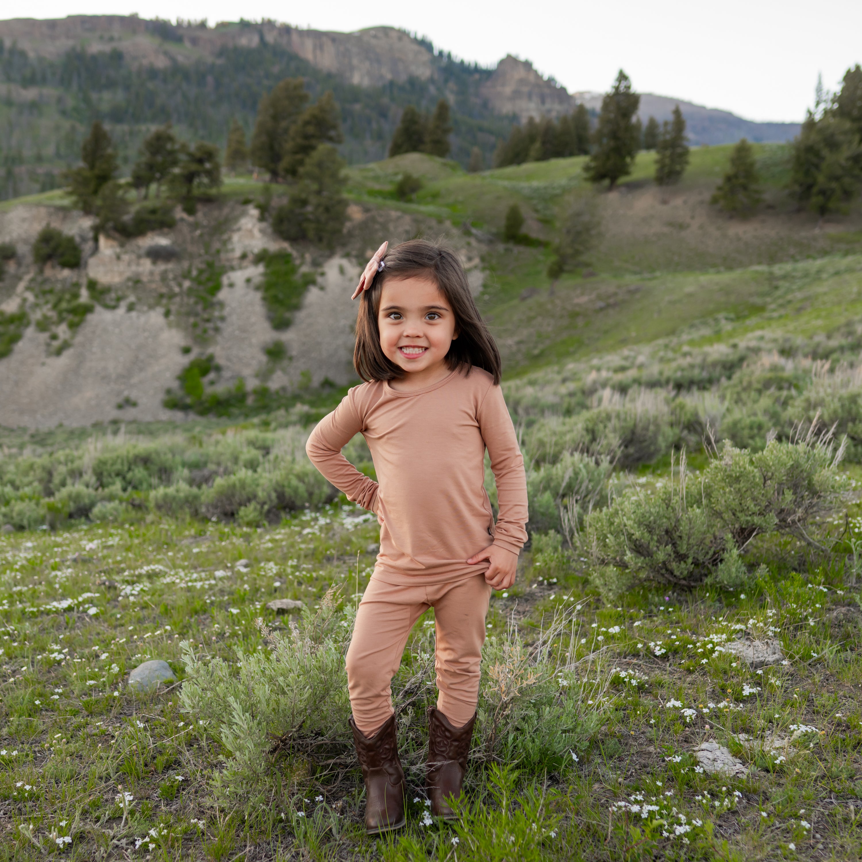 Girl model standing in the field with one hand on her hip wearing the Long Sleeve Pajamas in Latte and cowboy boots