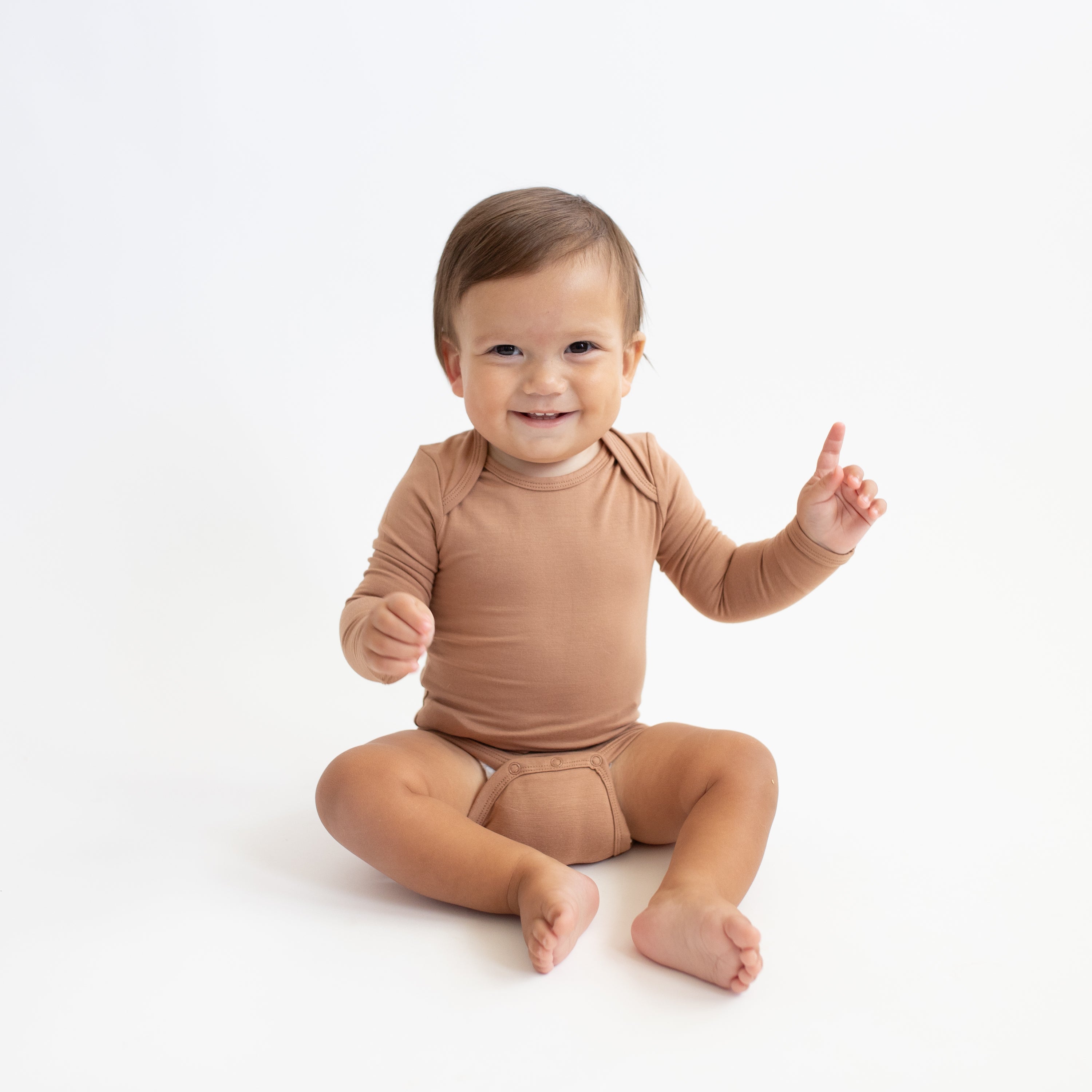 Smiling toddler sitting pointing upwards with one hand wearing the Long Sleeve Bodysuit in Latte