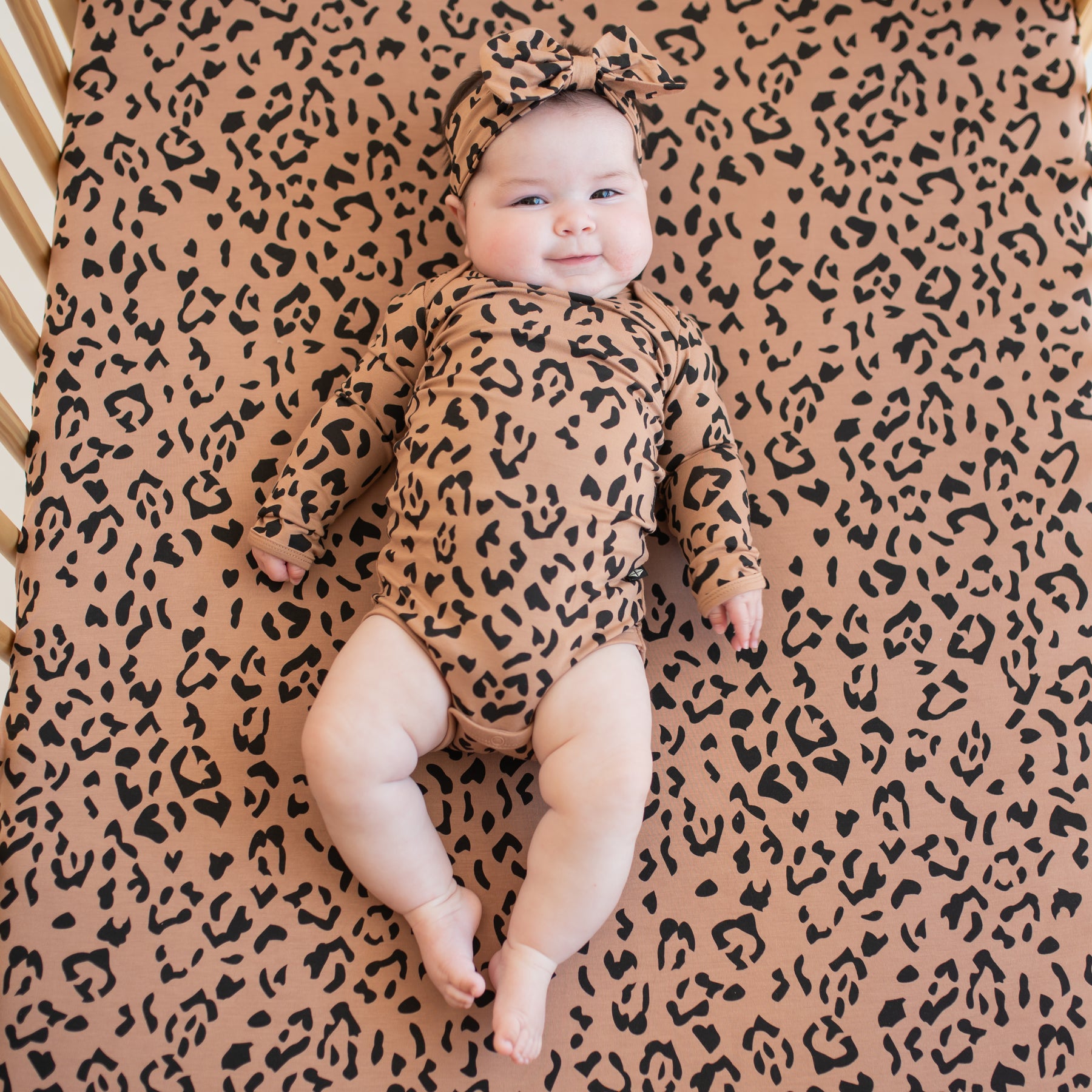 Infant wearing the Long Sleeve Bodysuit in Latte Leopard wearing a matching bow headband and laying on a matching crib sheet