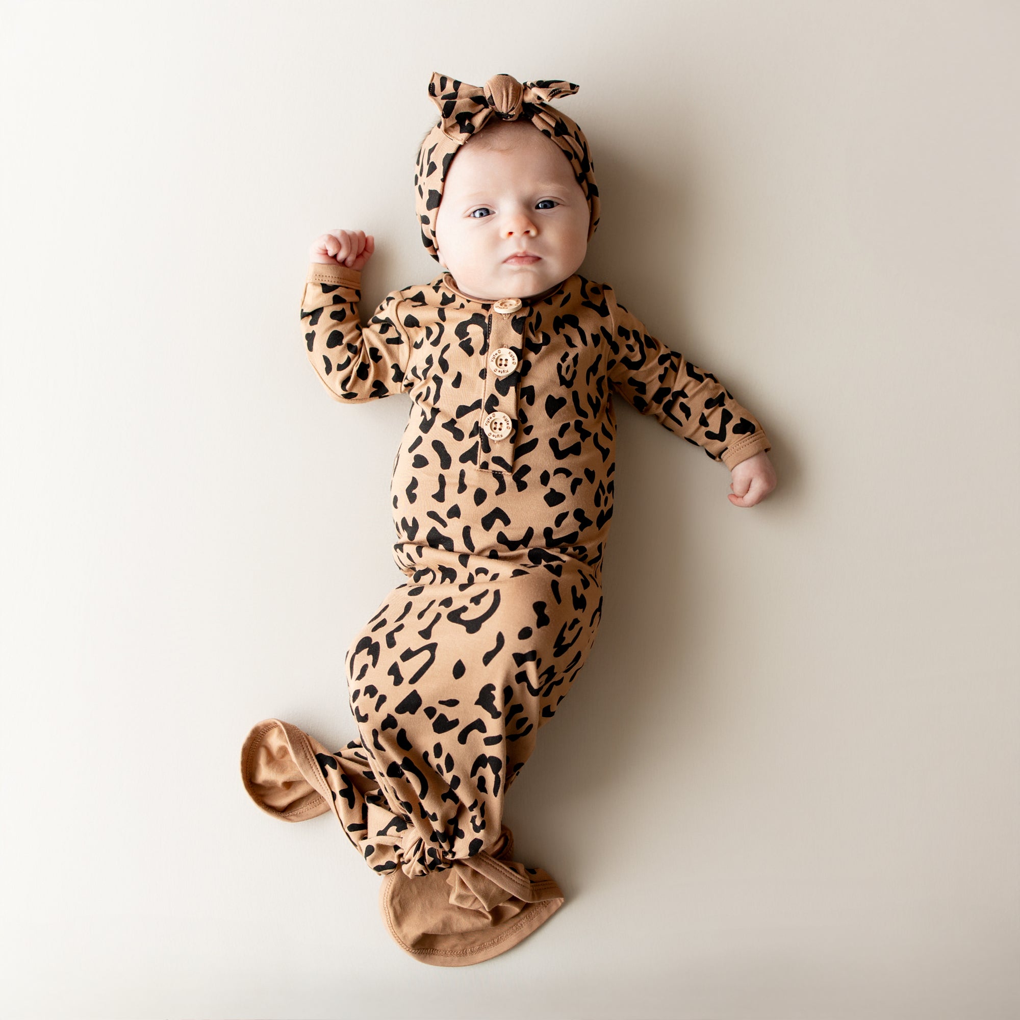 Infant laying on a neutral backdrop wearing the Knotted Gown with Bow Set in Latte Leopard
