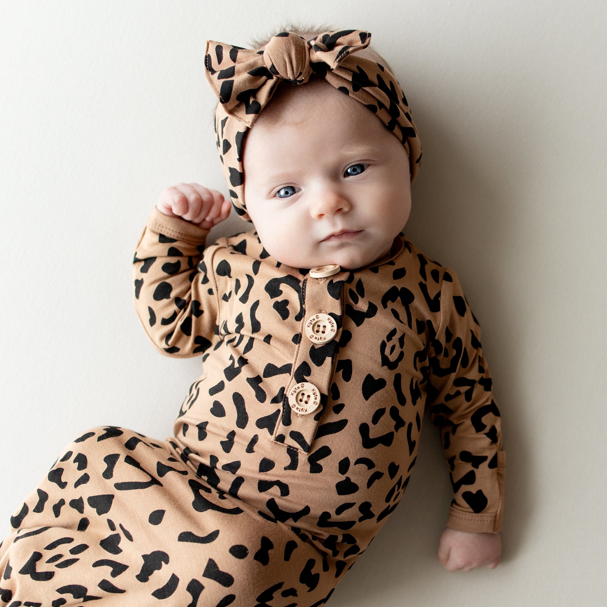 Infant laying on a neutral backdrop wearing the Knotted Gown with Bow Set in Latte Leopard