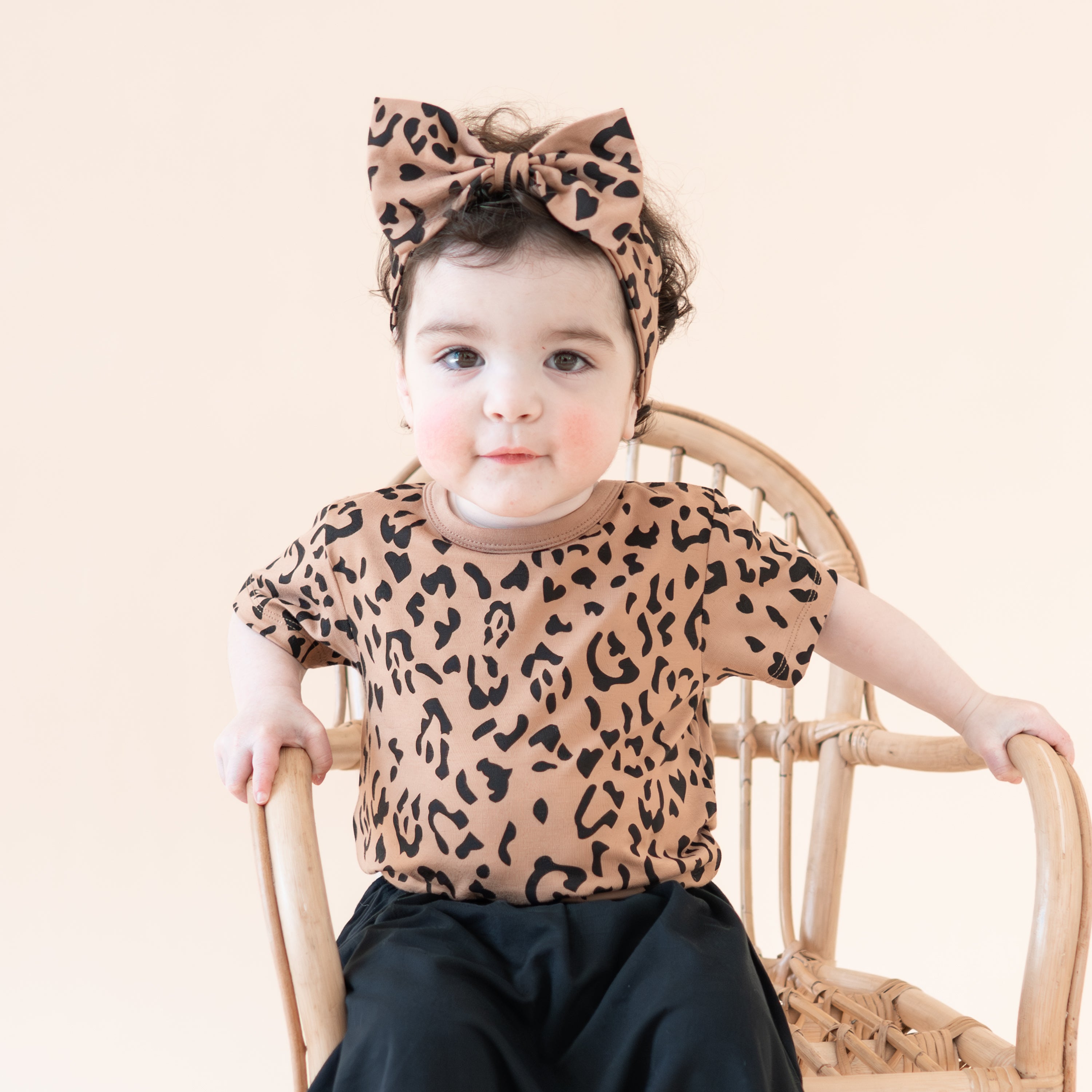Toddler sitting on a wicker chair earing the Toddler Basic Tee in Latte Leopard and matching bow with midnight skort