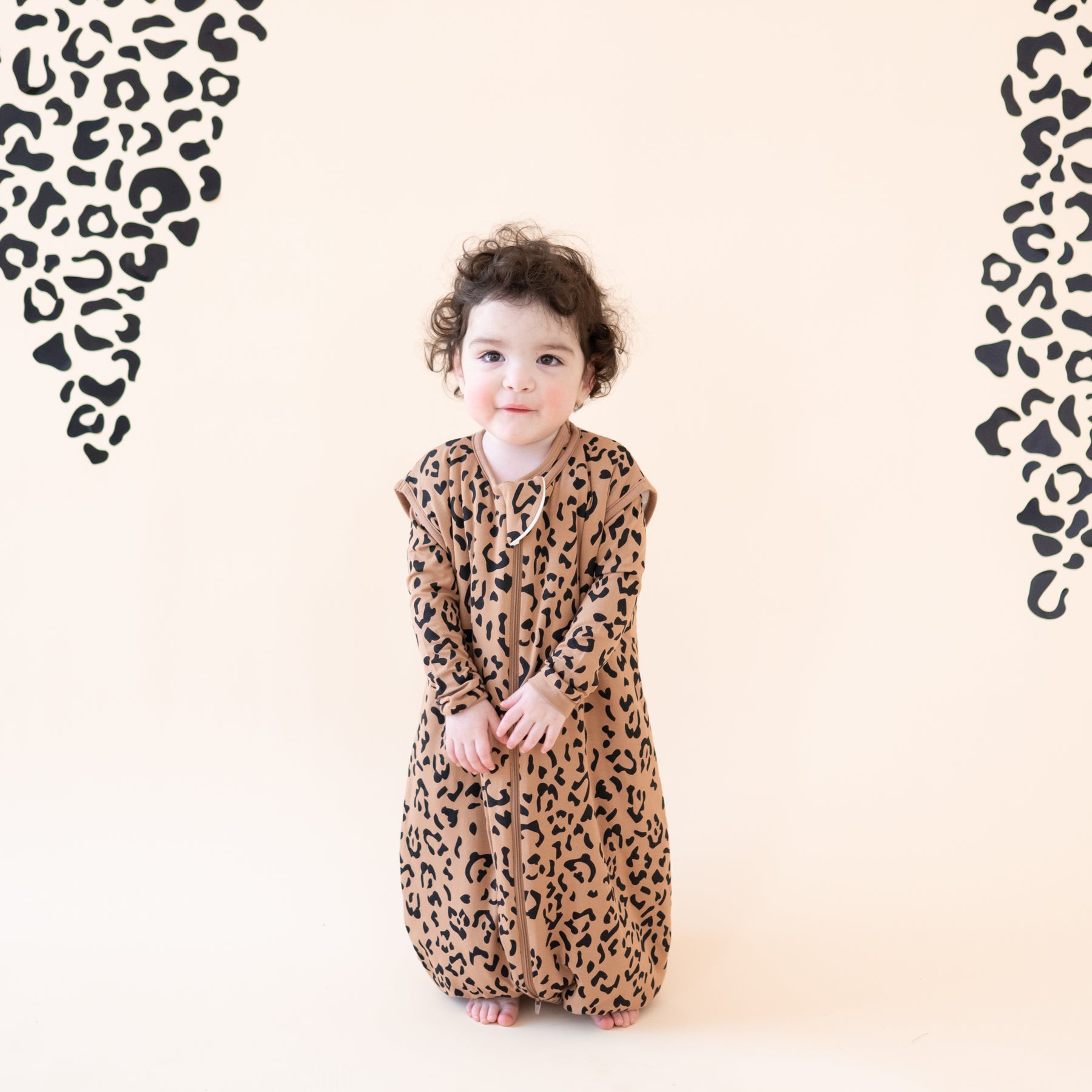 Toddler standing in front of a light colored leopard print backdrop wearing the Sleep Bag Walker in Latte Leopard 1.0