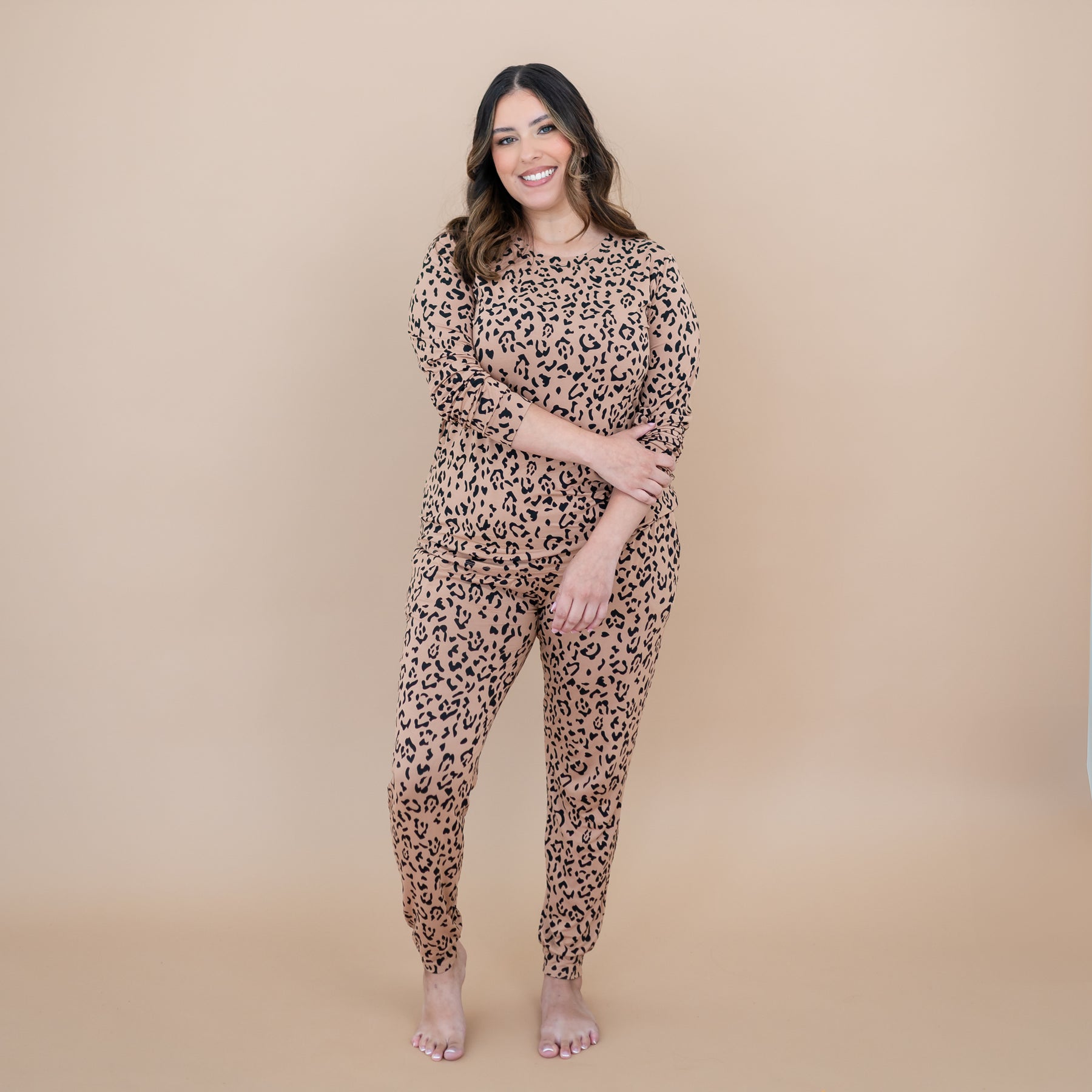 Smiling model standing with one hand holding the opposite arm wearing the Women's Jogger Pajama Set in Latte Leopard in front of a taupe background