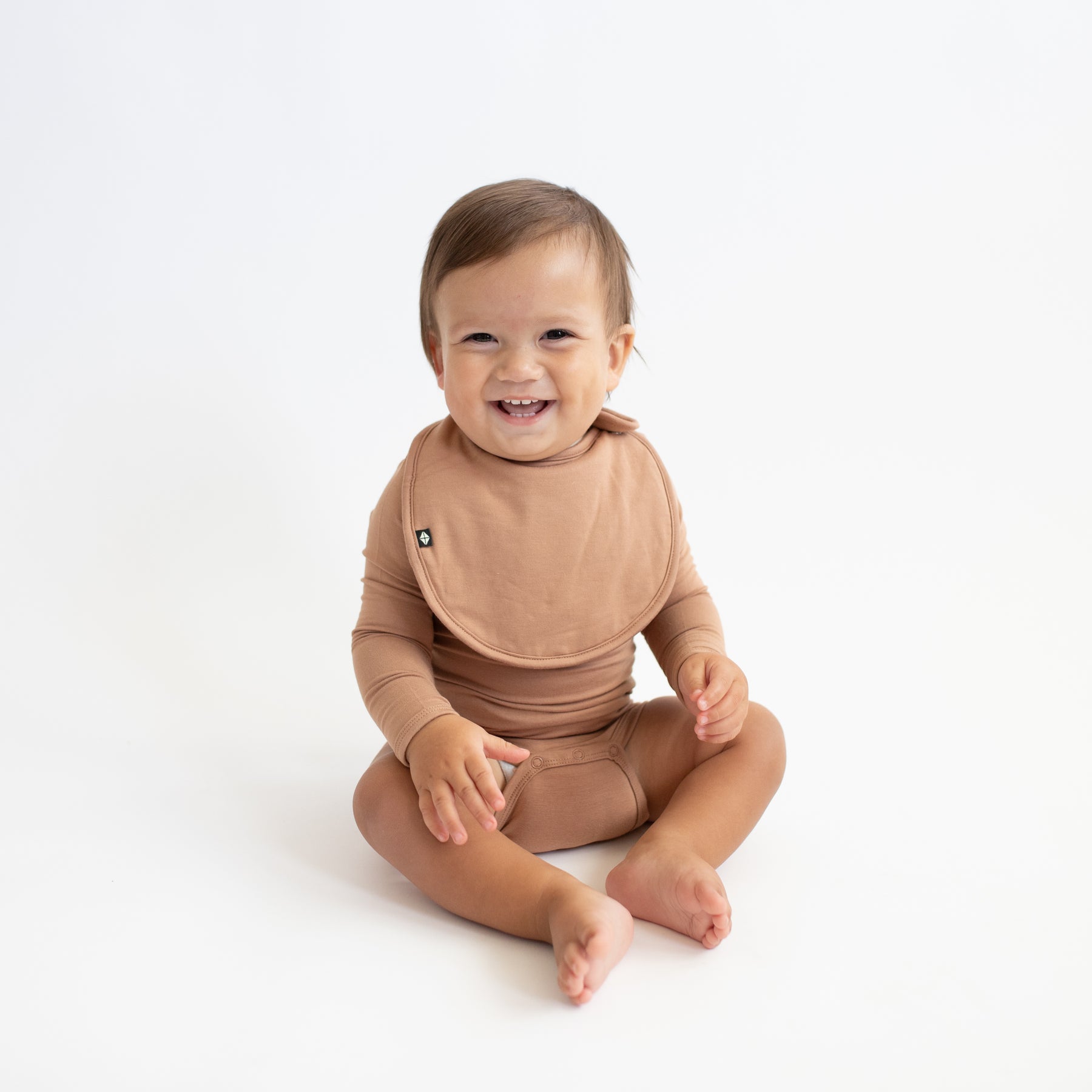 Smiling infant sitting wearing the Cushy Bib in Latte paired with a matching long sleeve bodysuit