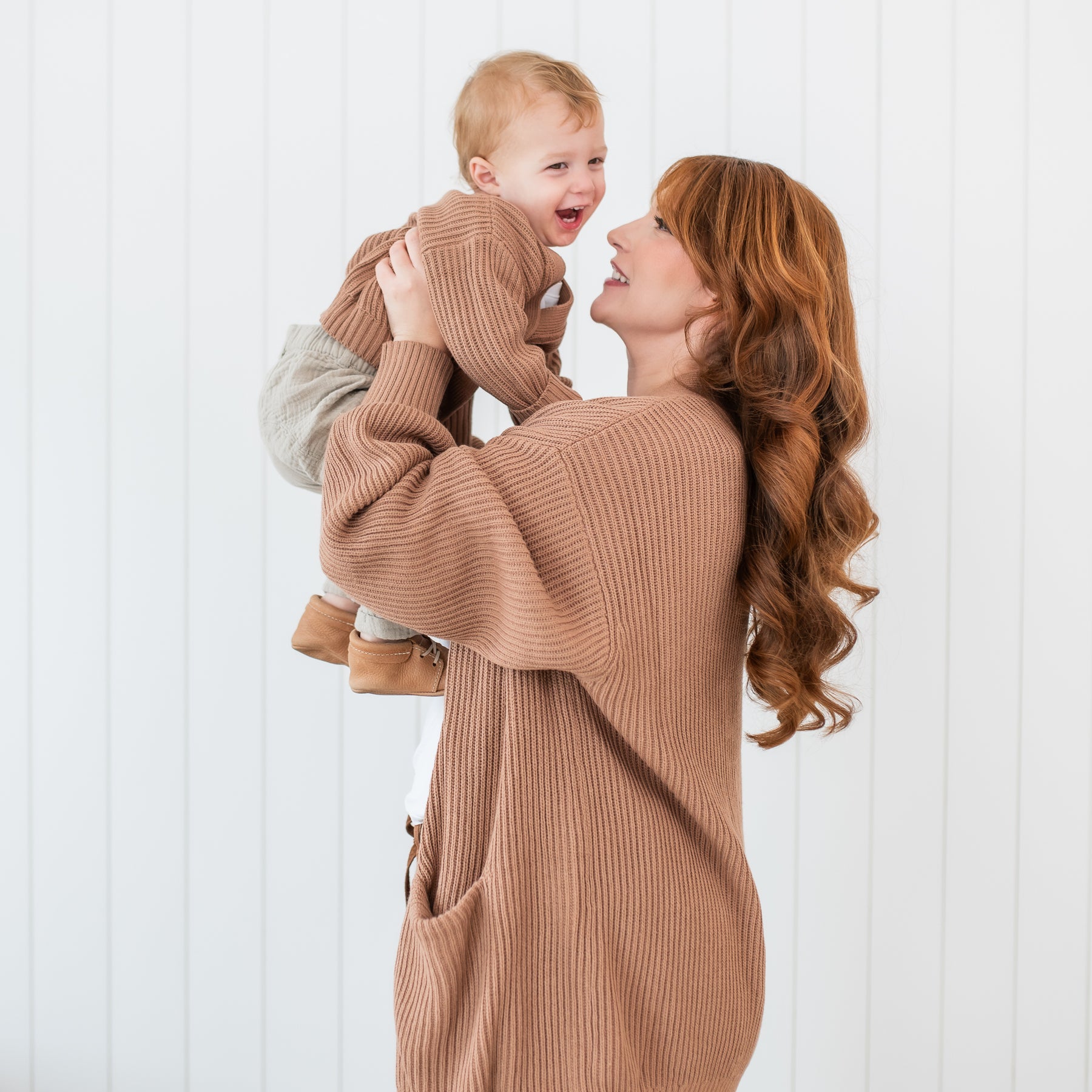 Mother wearing the Chunky Knit Women's Oversized Cardigan in Latte holding her son up looking at him