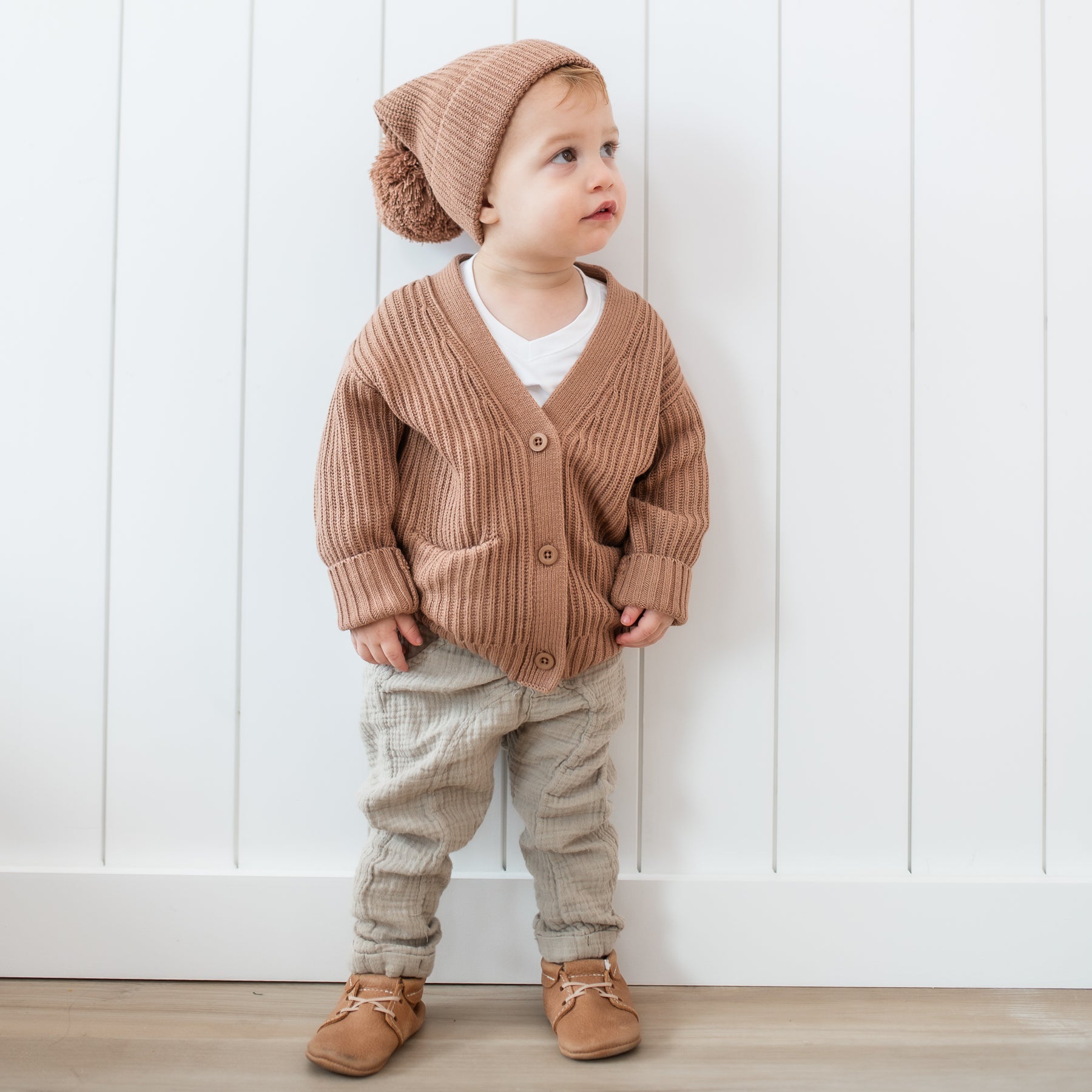 Toddler standing in front of a white paneled wall wearing the Chunky Knit Oversized Cardigan in Latte and matching chunky knit pom beanie