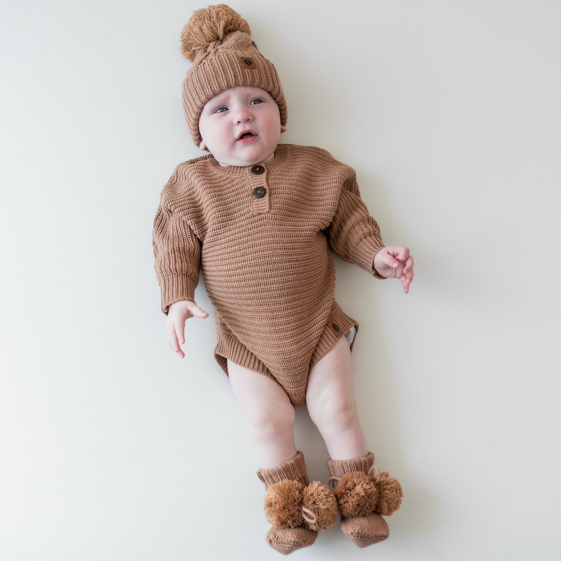 Infant laying down wearing the Chunky Knit Romper in Latte with matching knit booties and chunky cable knit beanie