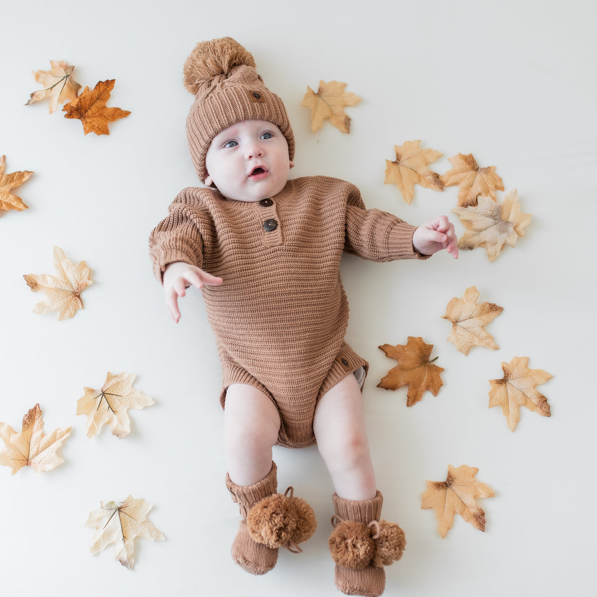 Infant wearing the Chunky Knit Romper in Latte with matching knit booties and chunky cable knit beanie surrounded by prop leaves
