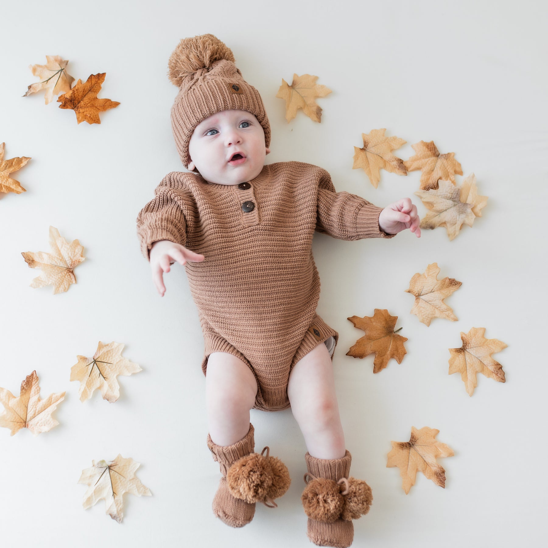 Infant wearing the Chunky Knit Romper in Latte with matching knit booties and chunky cable knit beanie surrounded by prop leaves