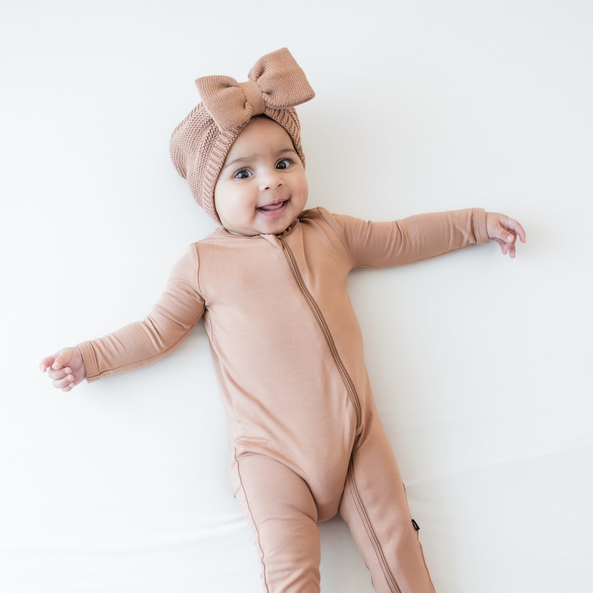 Infant laying down wearing the Chunky Knit Bow Headband in Latte paired with a matching zippered romper