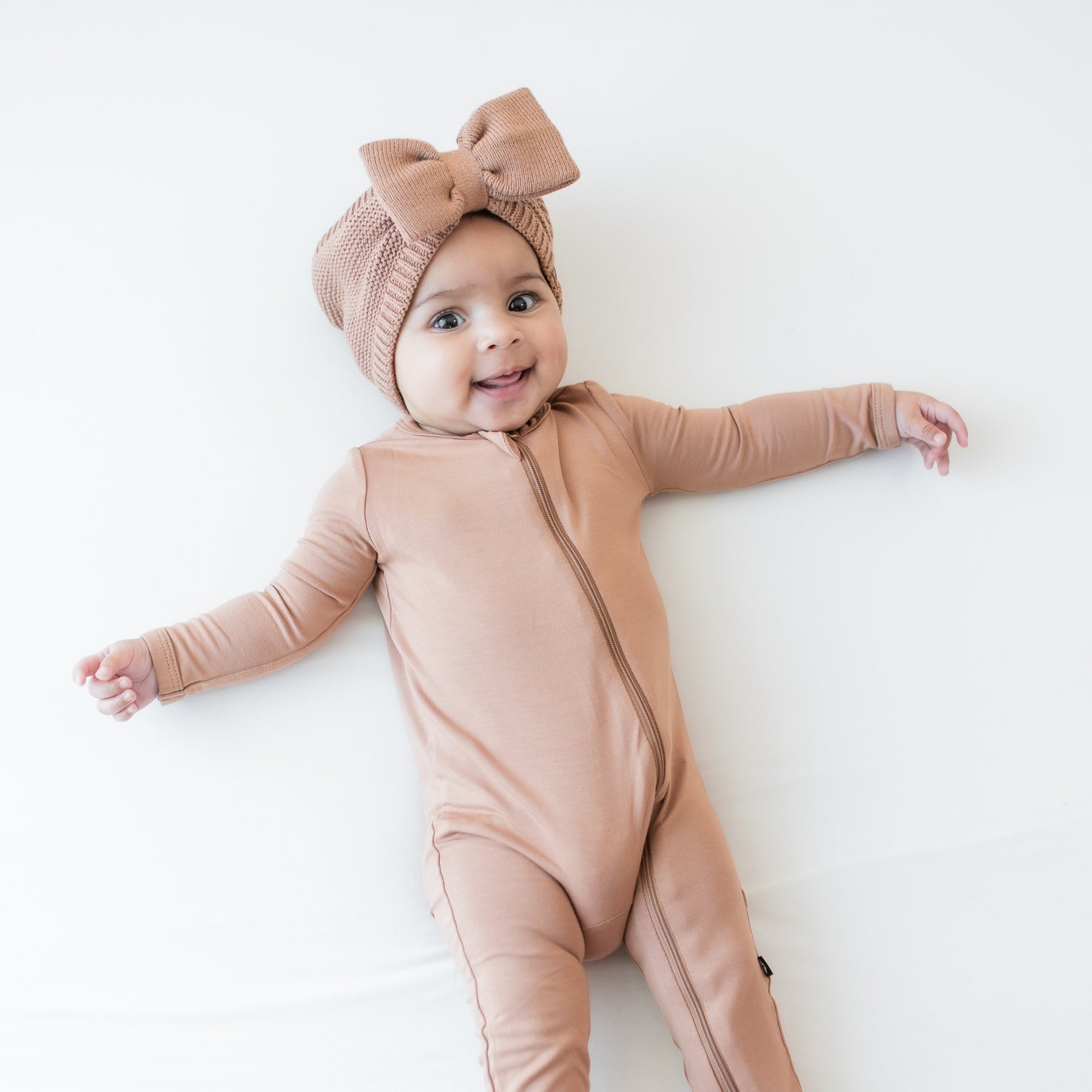 Infant laying down wearing the Chunky Knit Bow Headband in Latte paired with a matching zippered romper