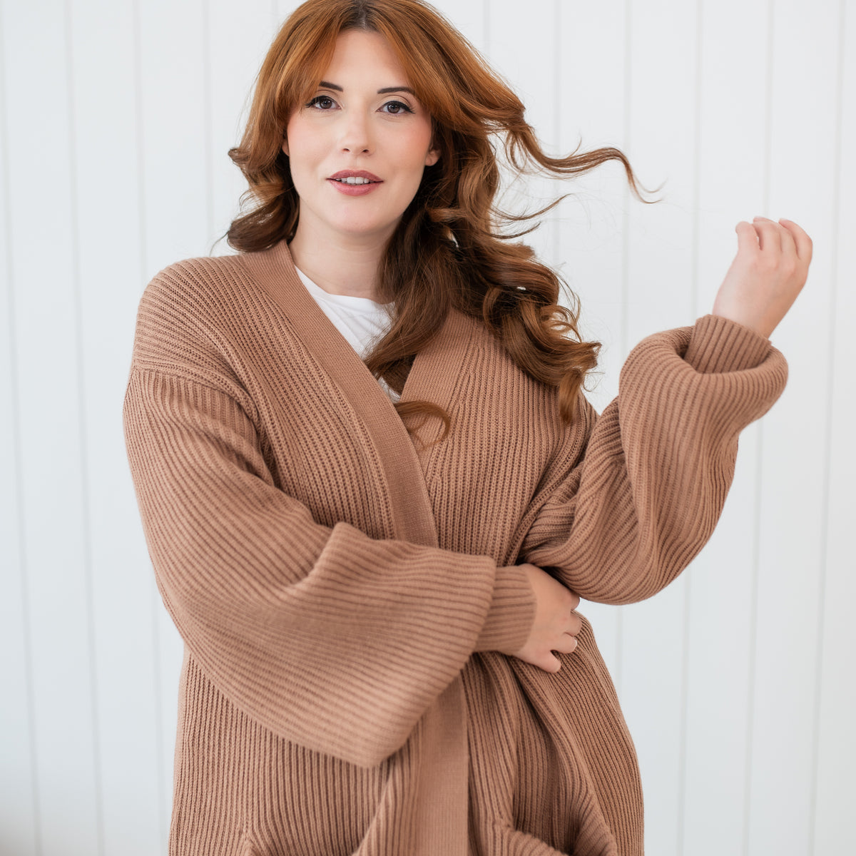 Model wearing the Chunky Knit Women's Oversized Cardigan in Latte standing in front of a white paneled wall