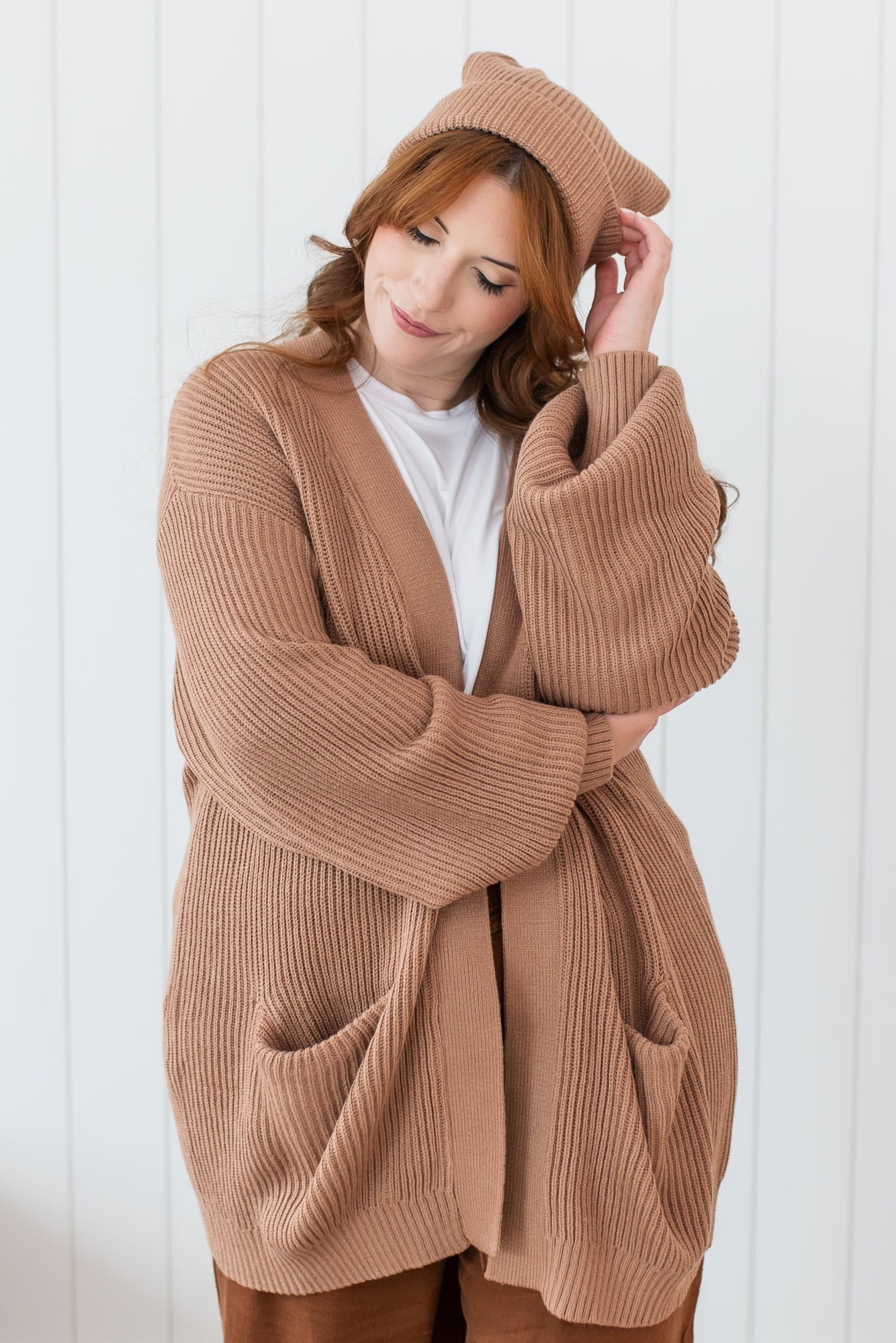 Female model standing in front of a white paneled wall wearing the Chunky Knit Women's Oversized Cardigan in Latte and matching chunky knit Adult pom beanie