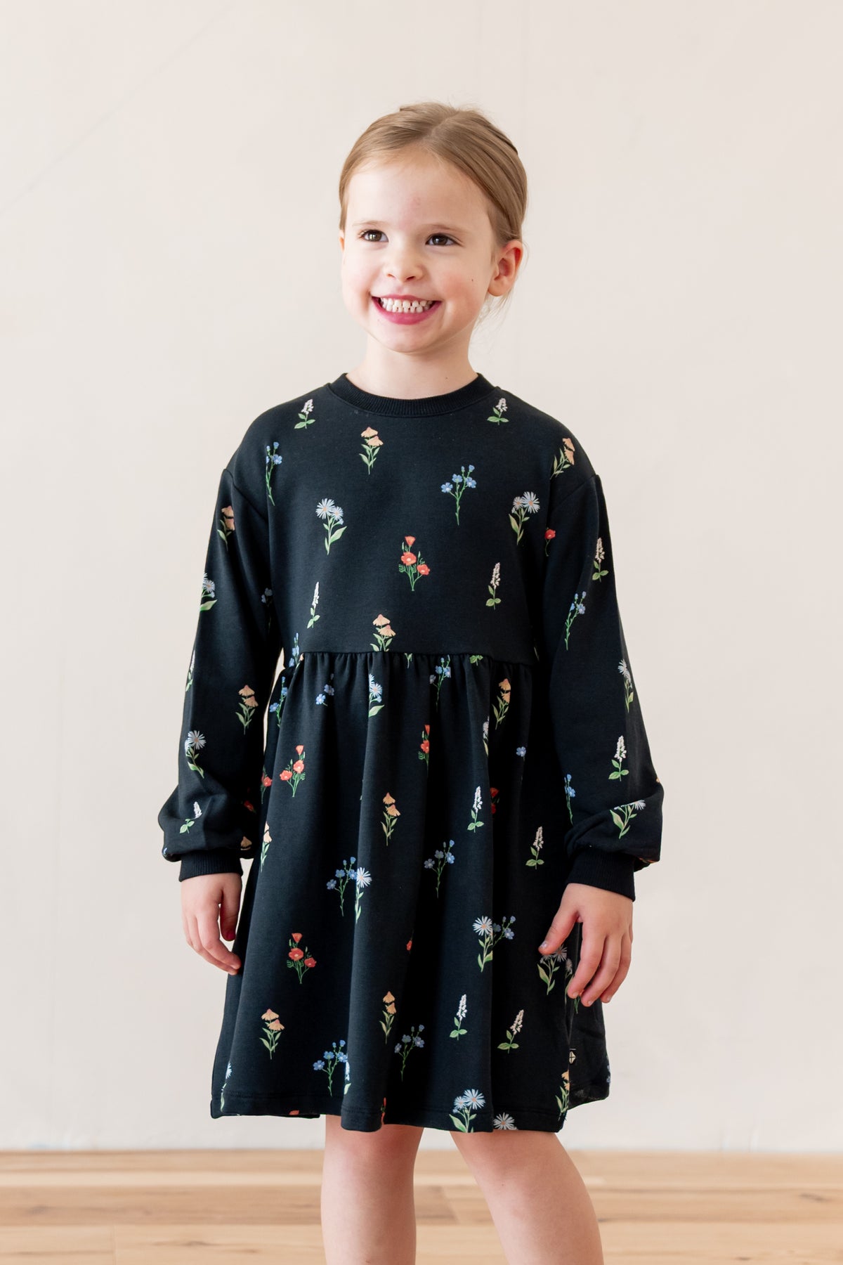 A young girl with blonde hair tied back is smiling and wearing a long-sleeve black dress with a colorful floral print, standing against a plain beige wall.