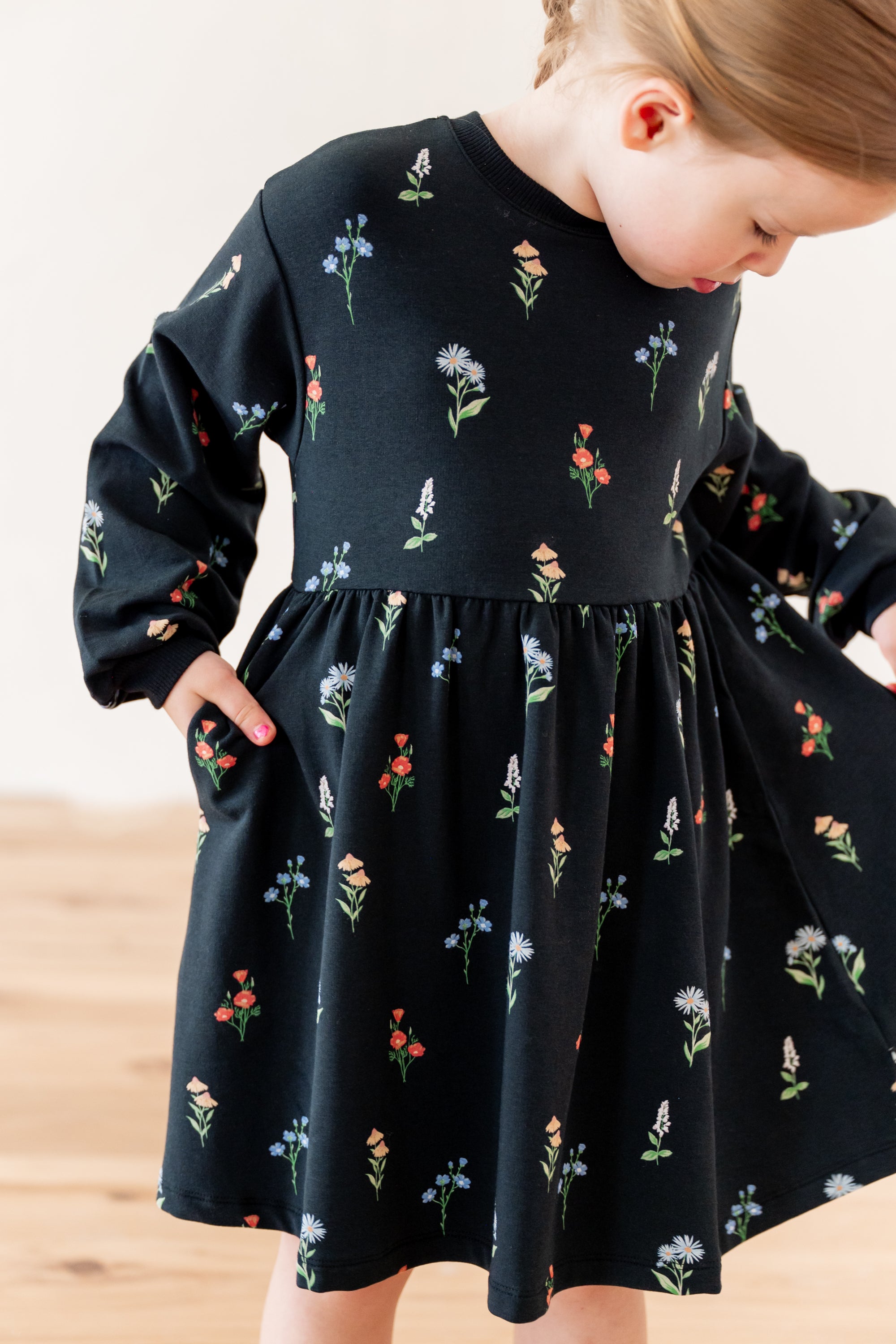 A toddler girl wearing a black long-sleeve dress with a colorful floral print with pockets, standing in a bright indoor setting.