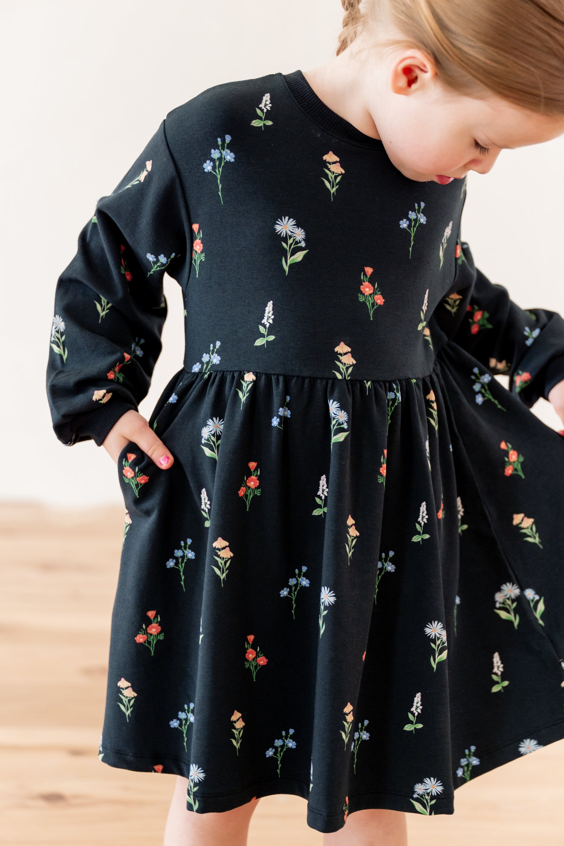 A toddler girl wearing a black long-sleeve dress with a colorful floral print with pockets, standing in a bright indoor setting.