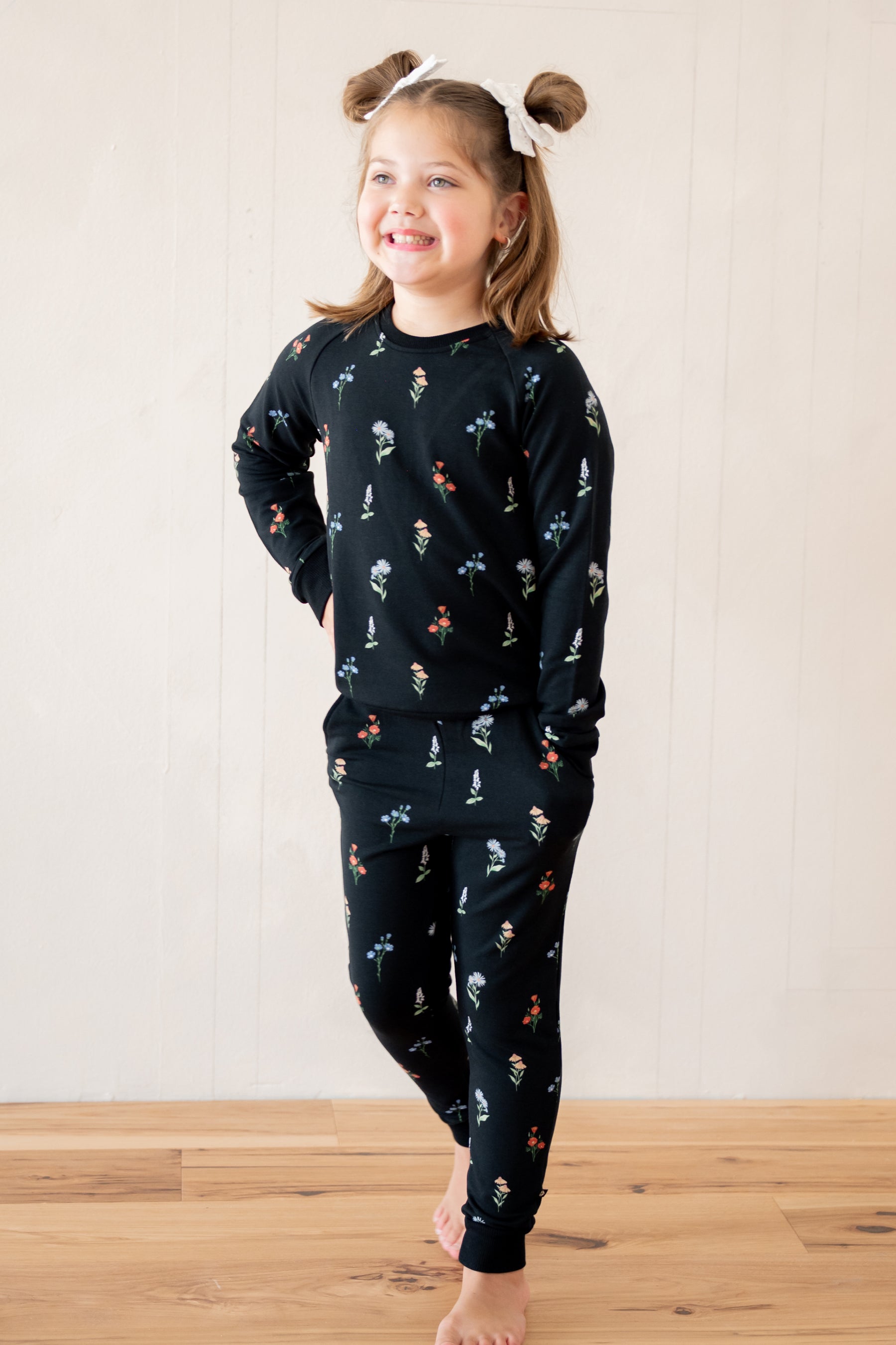 A young girl with brown pigtails wearing a matching black long-sleeve pajama set with a small multicolor winter pattern, standing barefoot on a wooden floor against a white paneled wall.