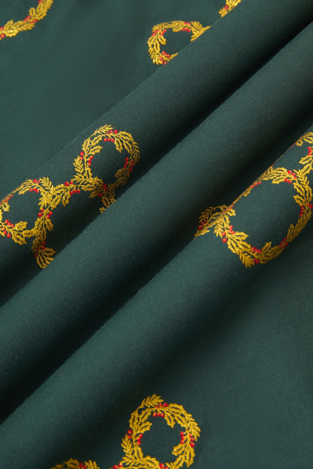 An all-over print of Mickey Mouse-shaped Christmas wreaths with gold and olive-green leaves and red holly berries, over a very dark green background color