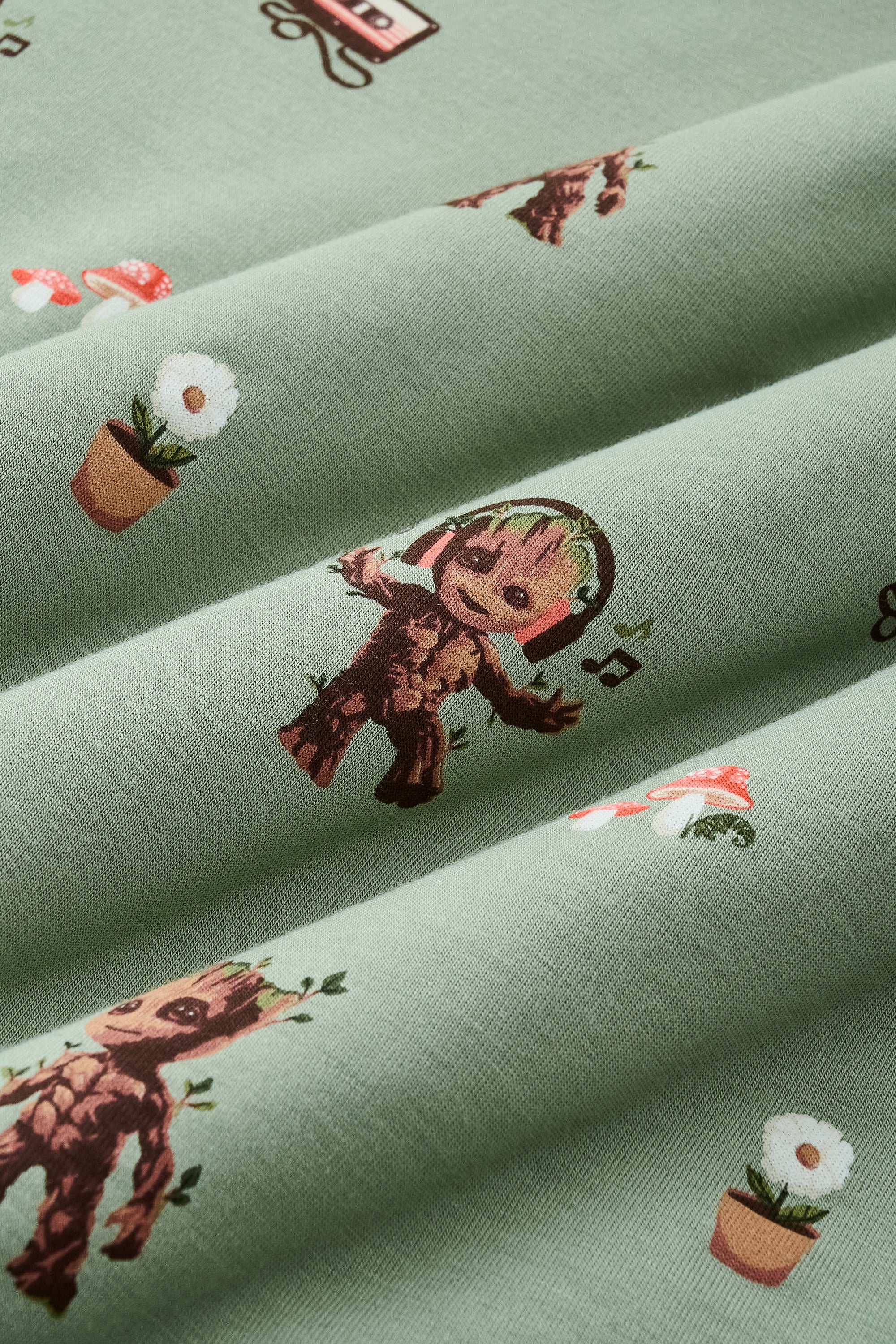 A repeating, all-over print featuring: a standing Baby Groot; a dancing Baby Groot wearing headphones and listening to music; a potted white daisy flower; two toadstools with red caps and white spots; a cluster of ivy; and a cassette tape with tape pulled out, over a dusty sage green background color named Thyme.